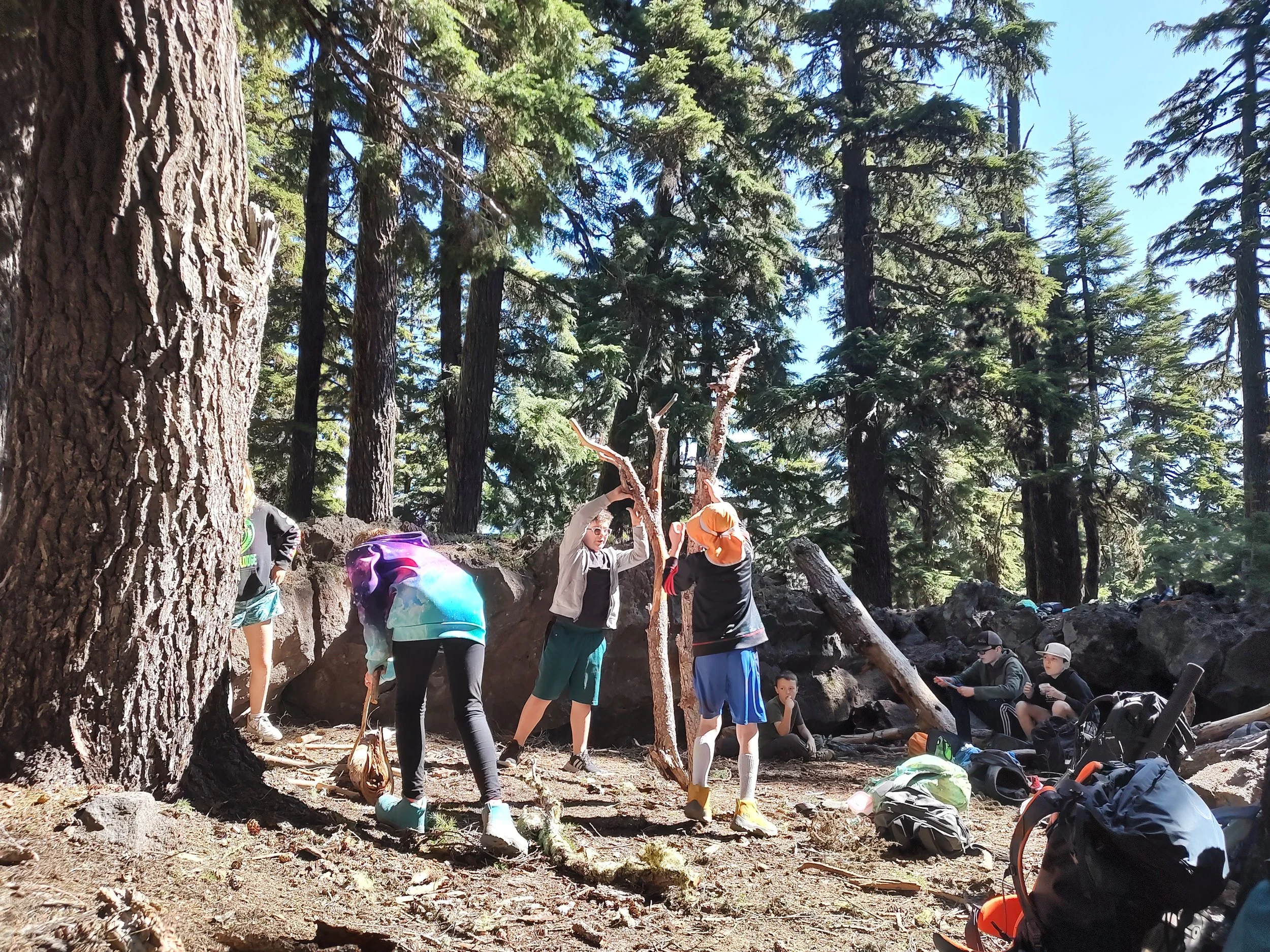 Group of children and teenagers in a forest, some sitting and some standing, with backpacks and outdoor gear, among tall trees and rocks, during daytime.