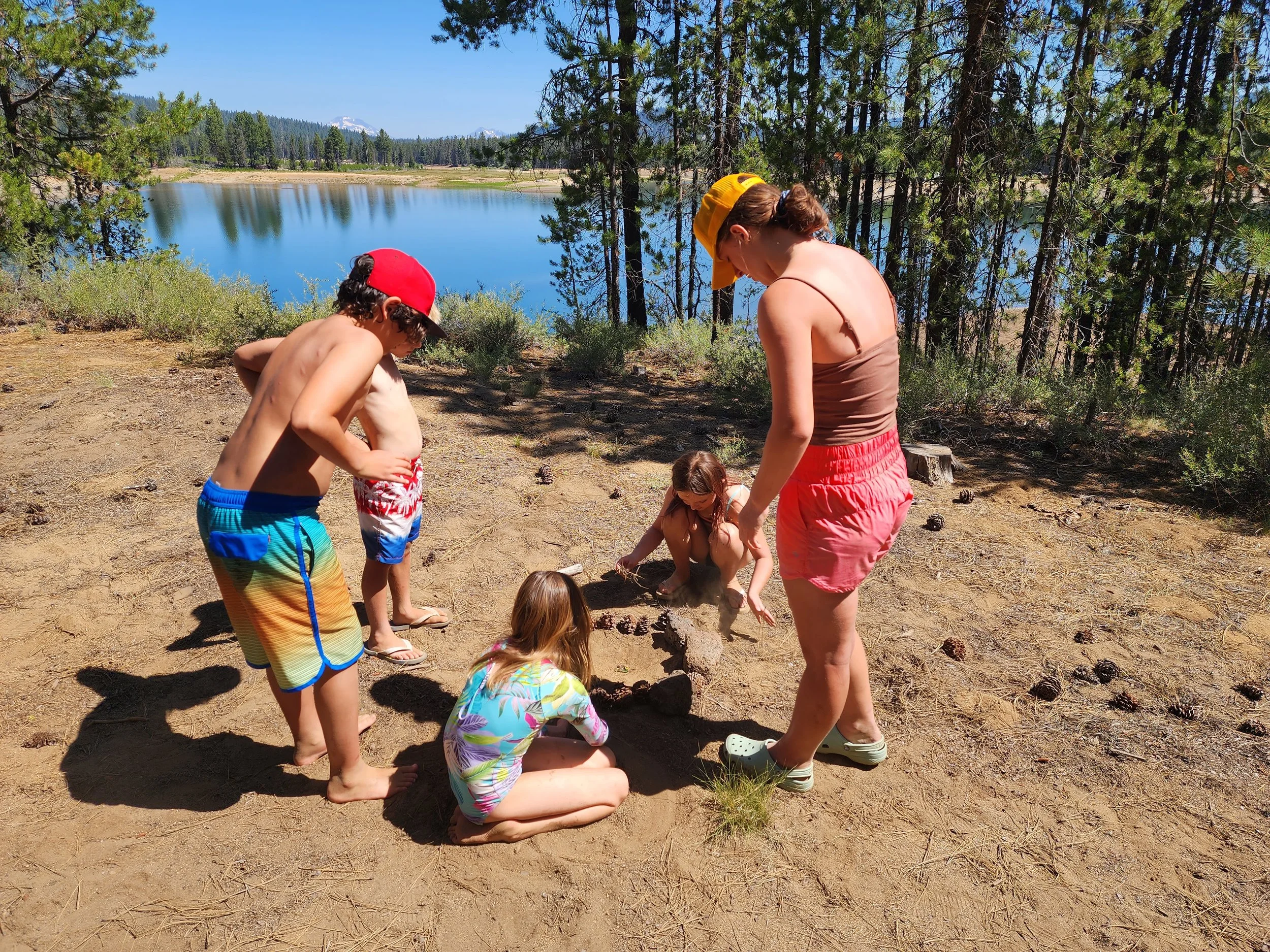 A woman and four children building a sandcastle near a lake with trees and mountains in the background on a sunny day.