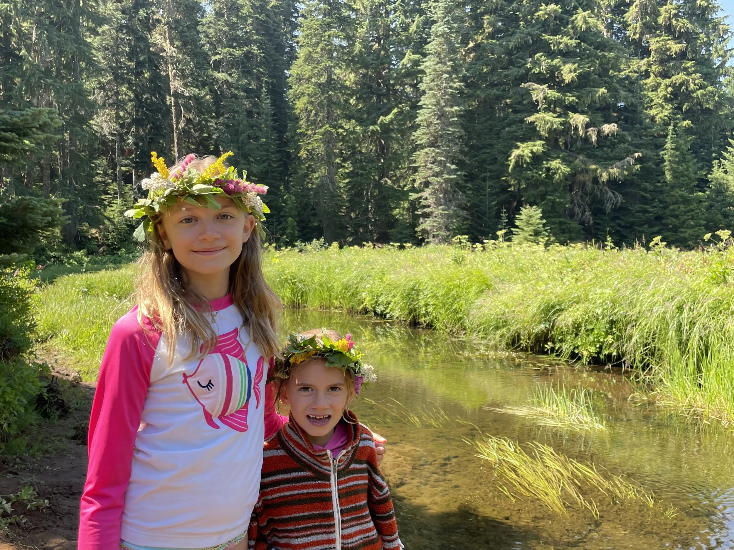 Two young girls wearing flower crowns stand by a small creek in a forested area, smiling at the camera.