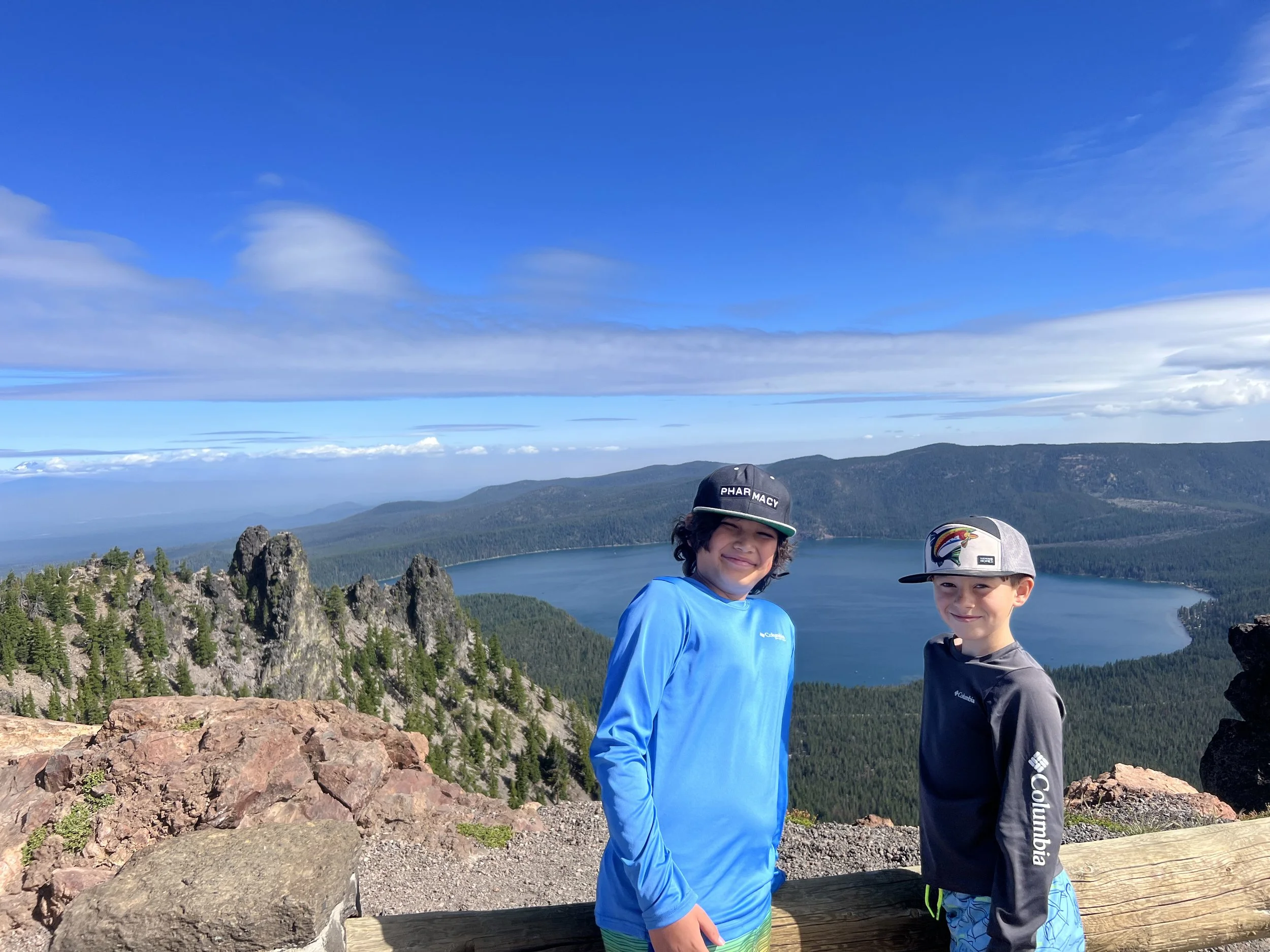 Two boys standing on a rocky overlook with a scenic mountain lake and forested hills in the background under a partly cloudy sky.