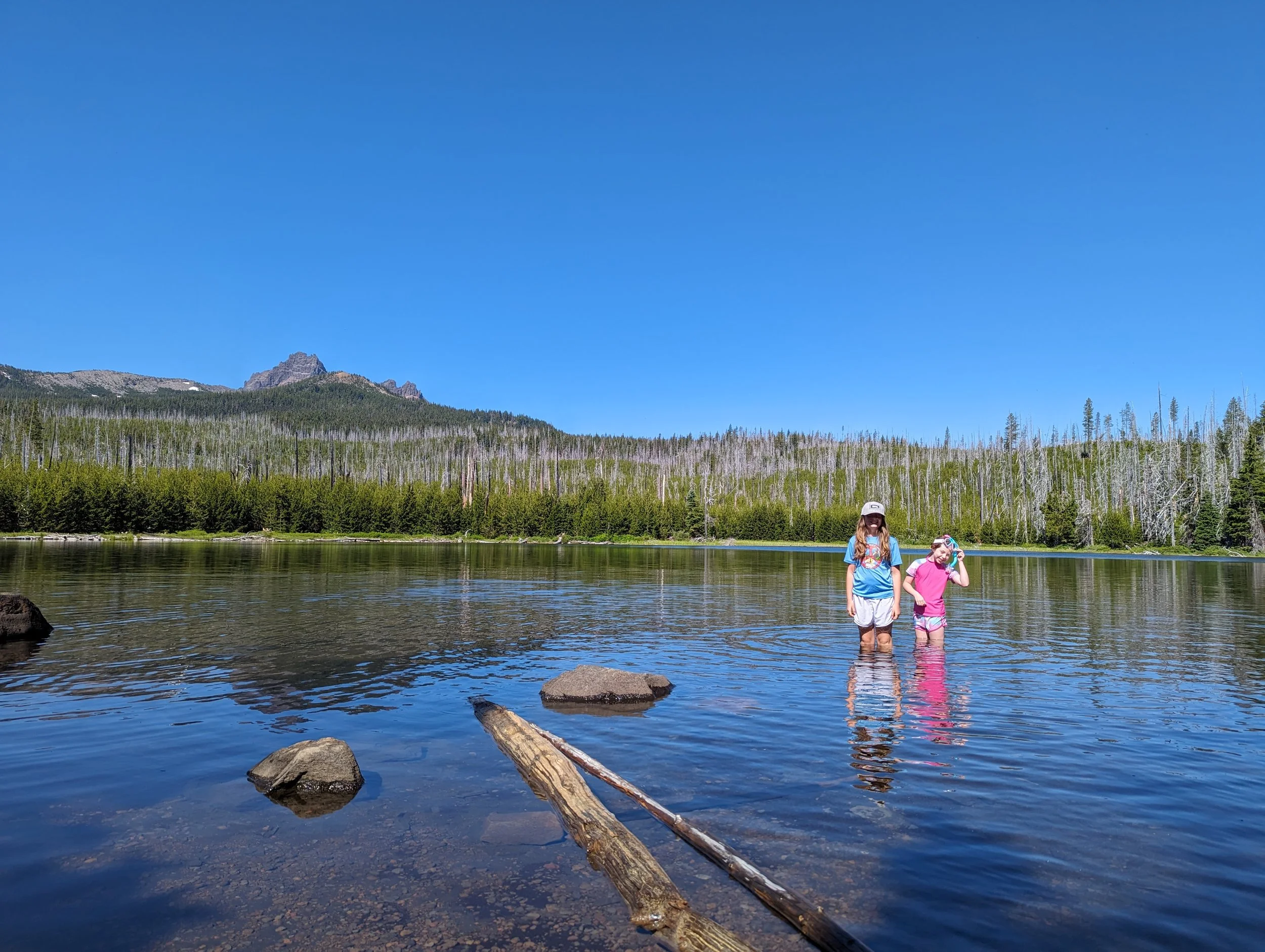 Two young girls standing in a calm river with a forest and mountain in the background, under a clear blue sky.