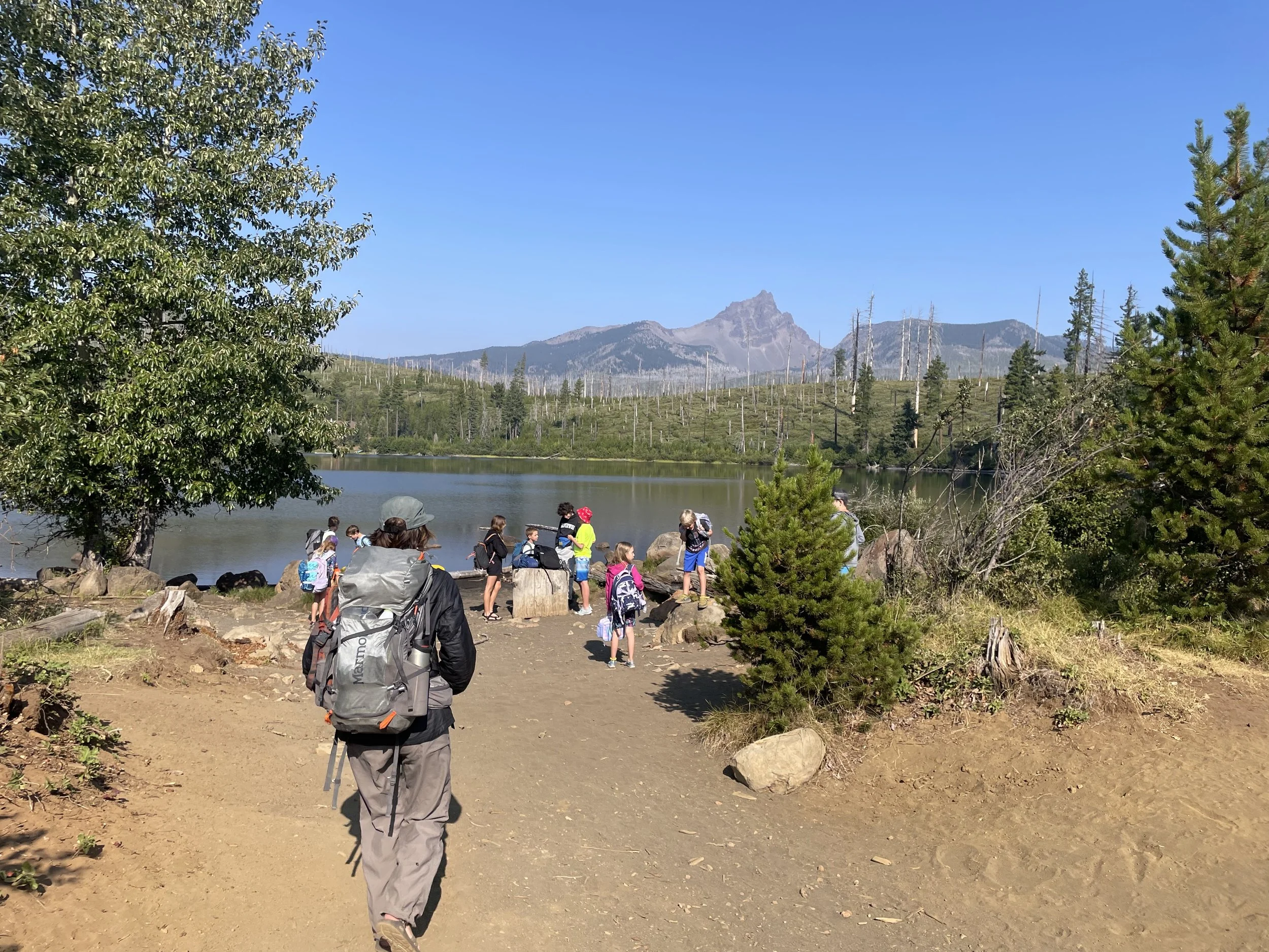 A group of children and adults on a hiking trip near a lake in a forest with mountains in the background.