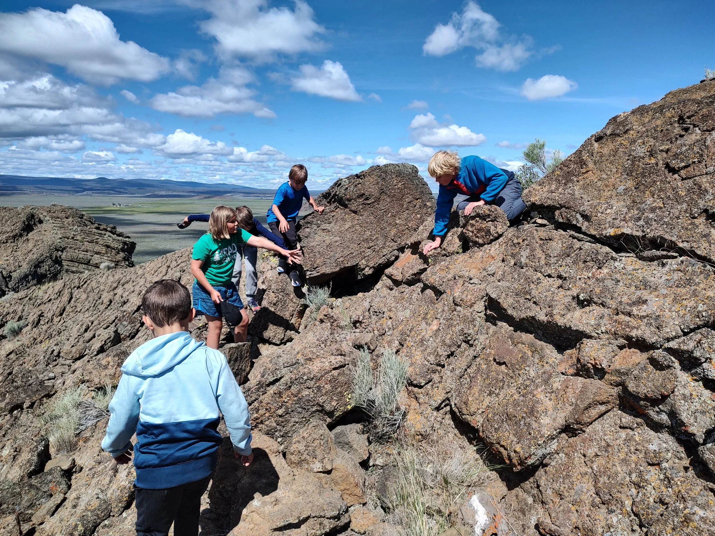 Children climbing on rocky terrain outdoors on a sunny day with blue skies and scattered clouds.