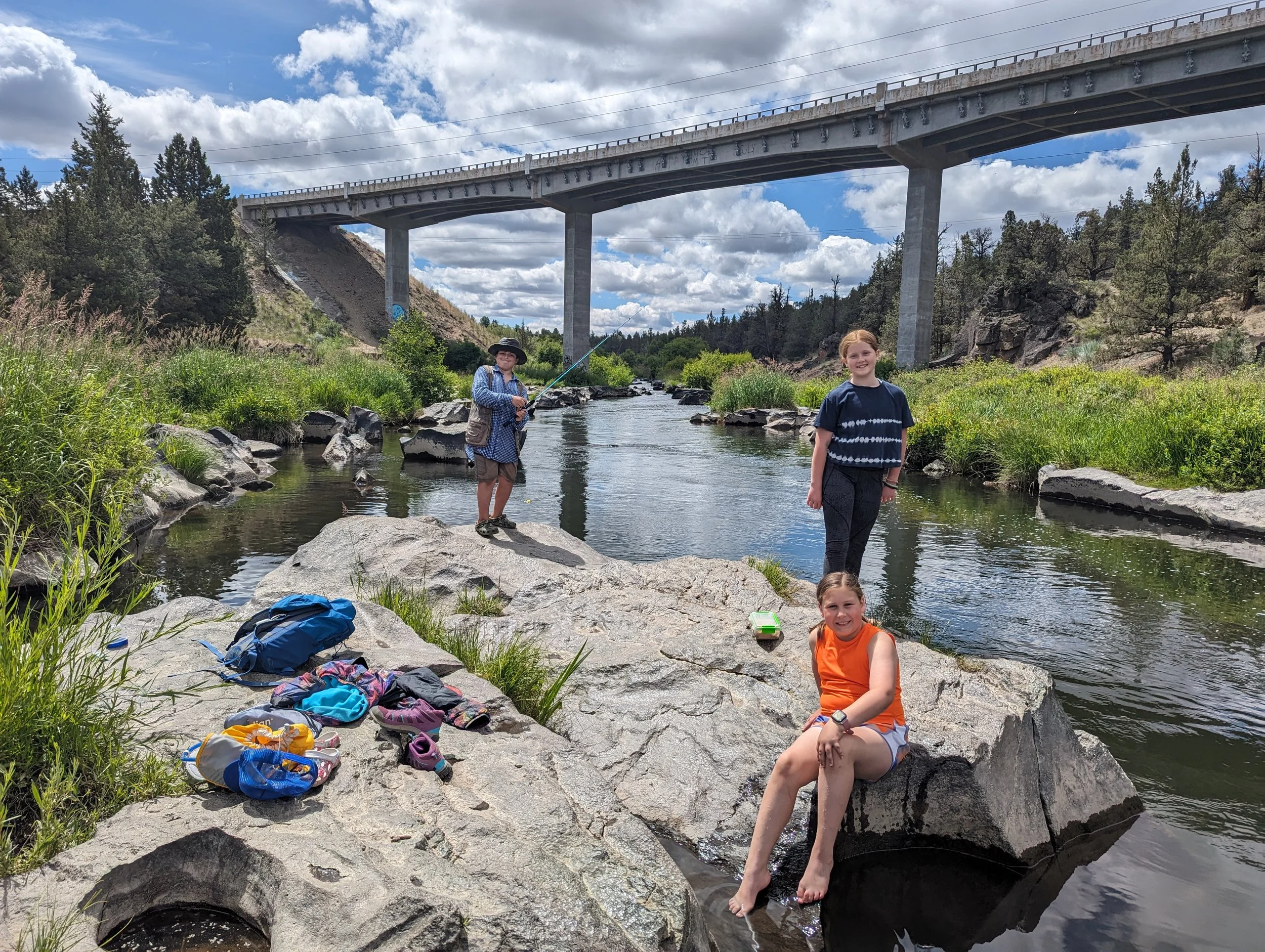Three children by a river, with one girl sitting on a rock and two girls standing near the water, one of whom is fishing, with a bridge overhead and a cloudy sky.