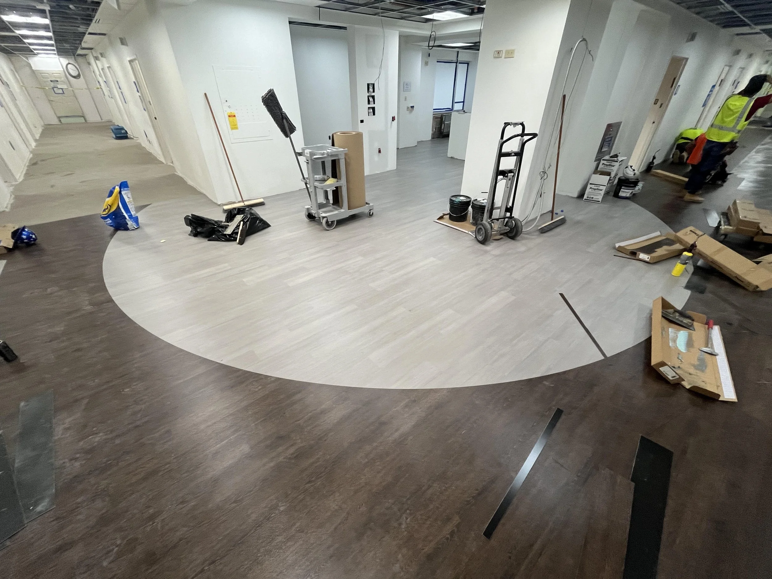 Office space under renovation with new light-colored wood flooring installed in a circular pattern, and construction tools and supplies scattered around.