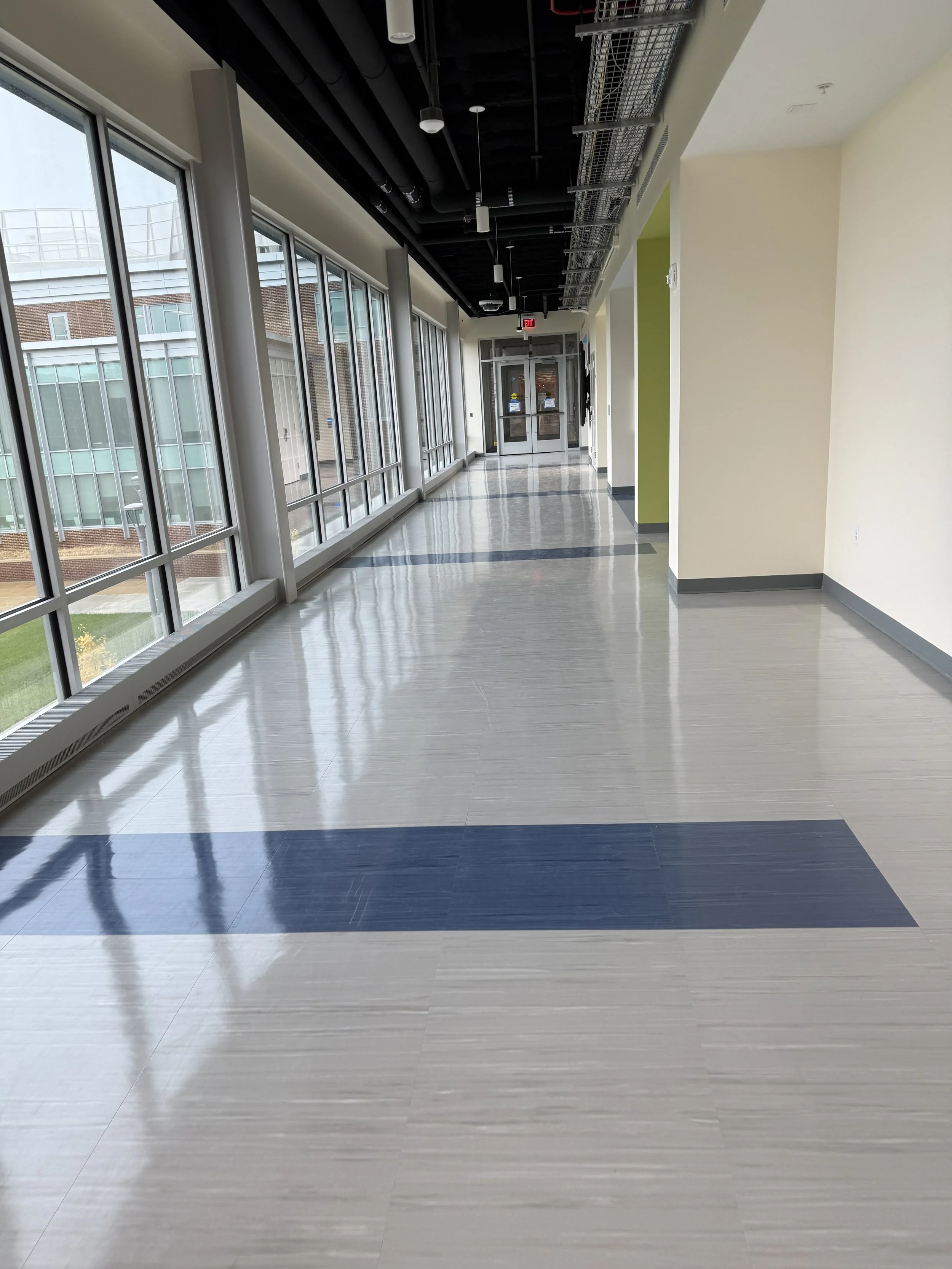 Empty corridor with large windows on the left, beige and green walls on the right, and a glossy tiled floor with dark and light strips, leading to glass doors at the end.