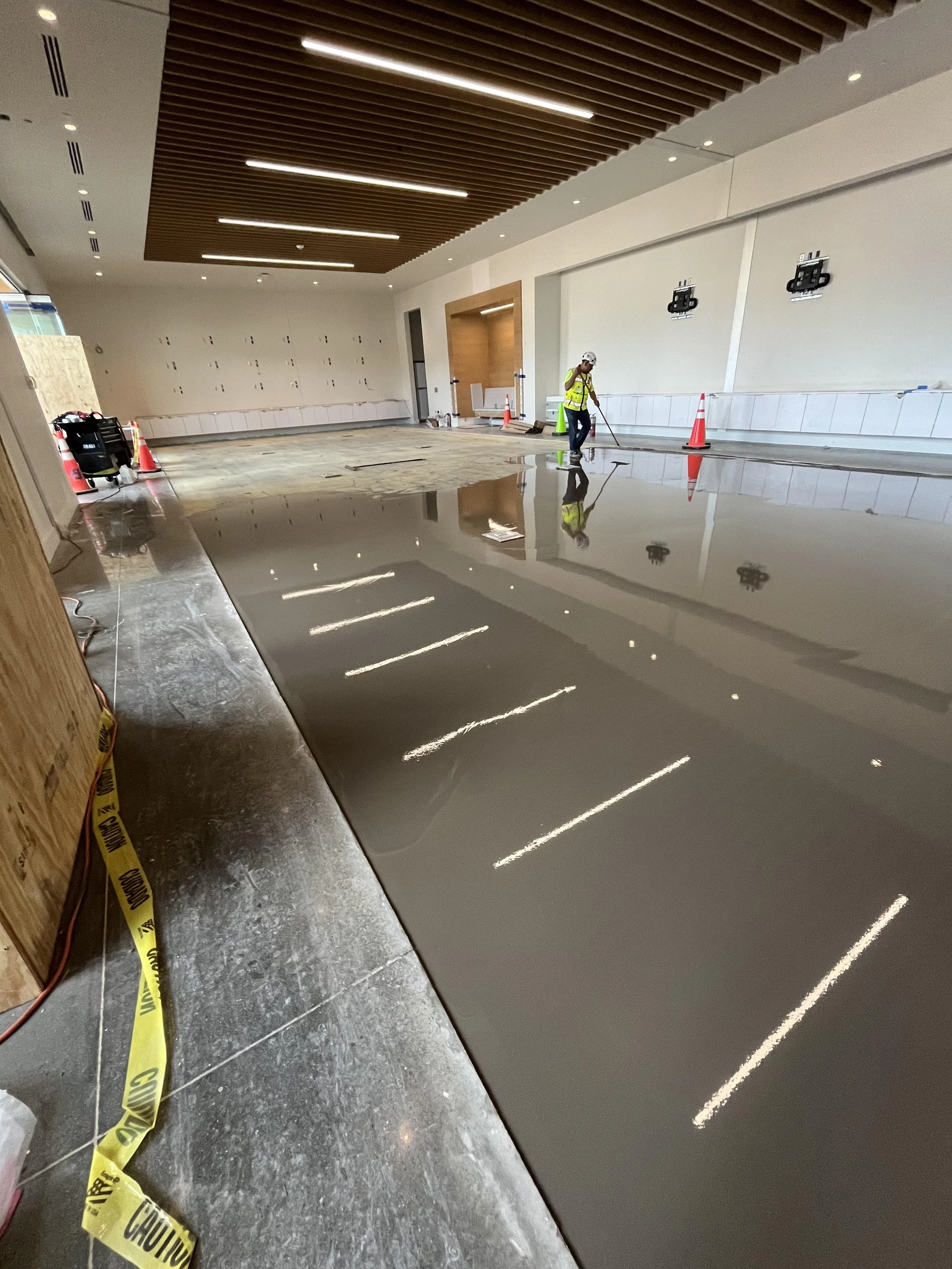 Construction worker in a yellow safety vest and hard hat polishing a shiny concrete floor inside a building with modern interior design and wooden ceiling panels.