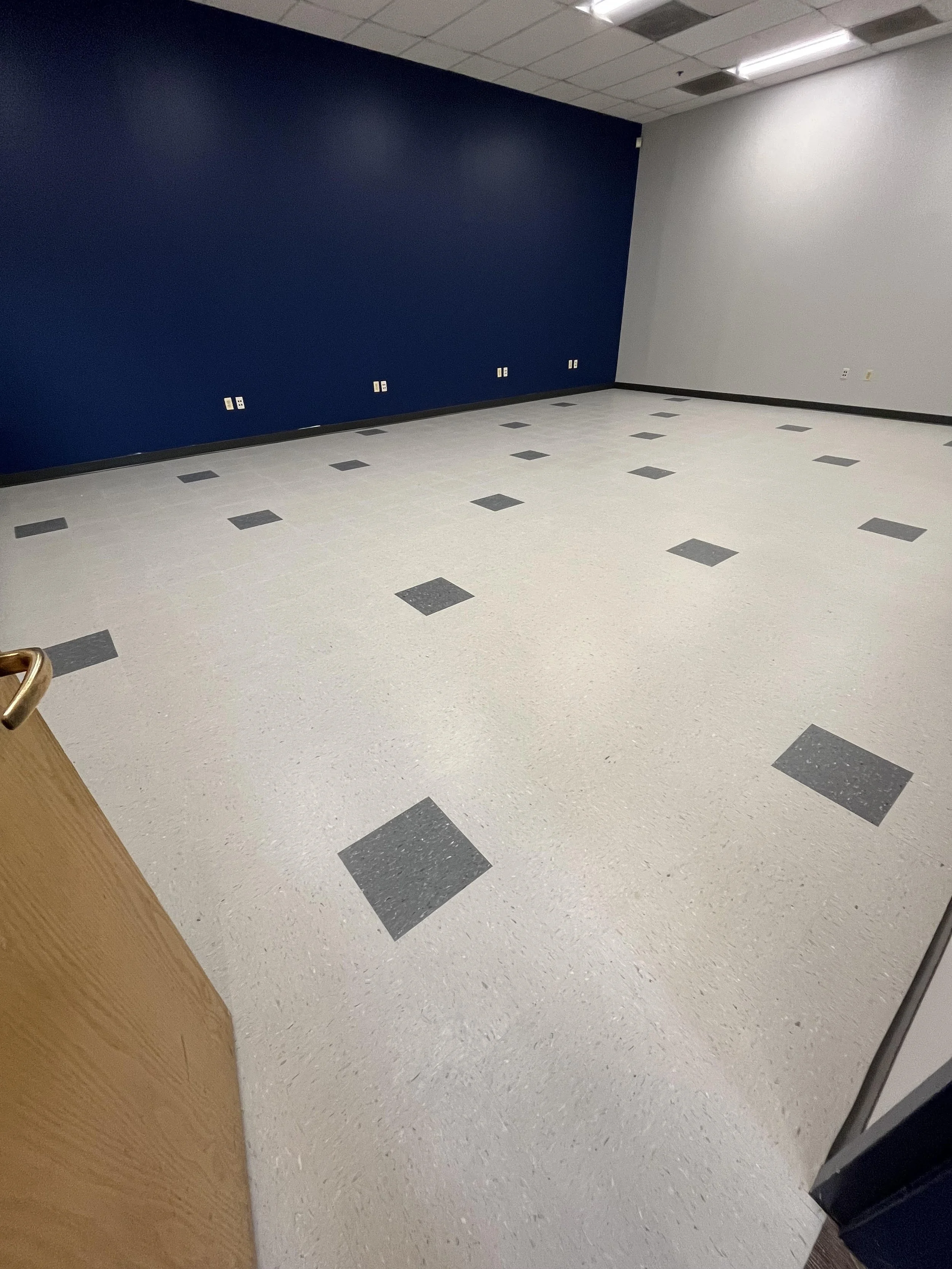 Empty room with white and blue walls, light-colored speckled tile floors with darker square tiles, and ceiling with fluorescent lights.
