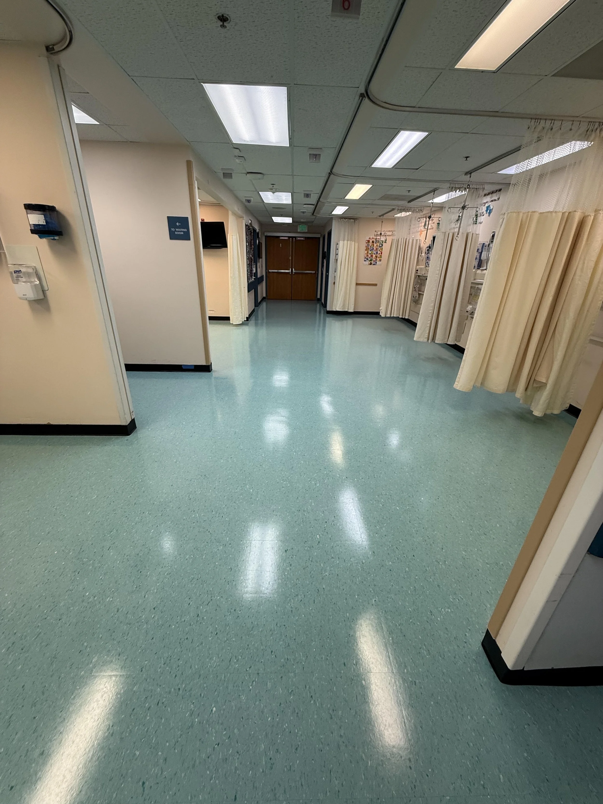 Hospital hallway with cream-colored walls, light green flooring, medical curtains on the right side, and ceiling lights.