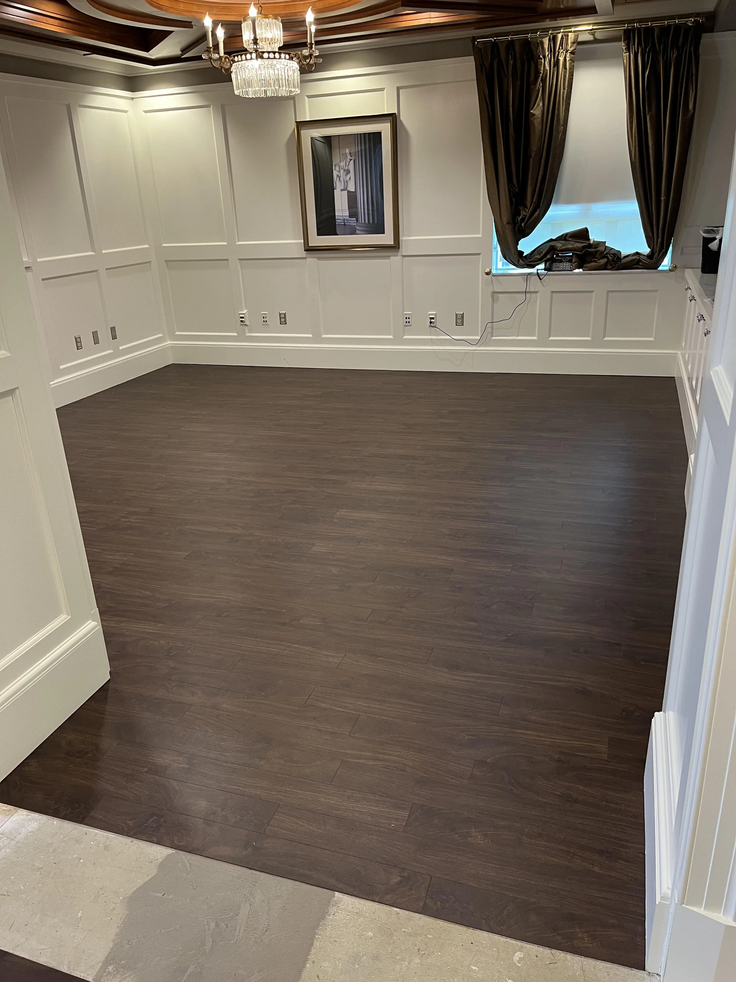Empty dining room with dark hardwood floors, white paneled walls, a framed picture, a window with dark curtains, and a chandelier ceiling light.