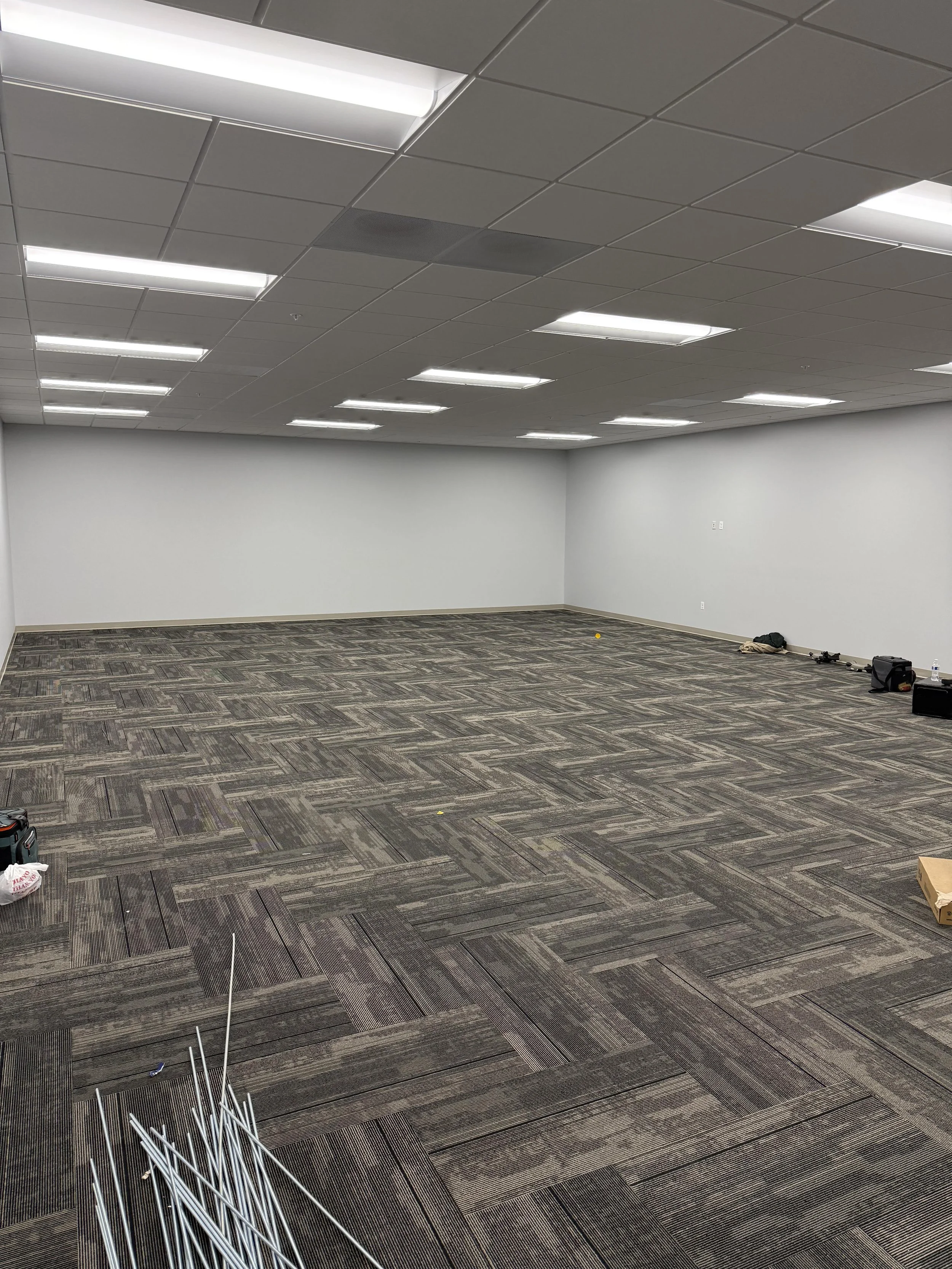 Empty room with white walls, gray ceiling tiles, modern lighting, and patterned gray carpet flooring. There are some tools and supplies on the floor, indicating ongoing setup or renovation.