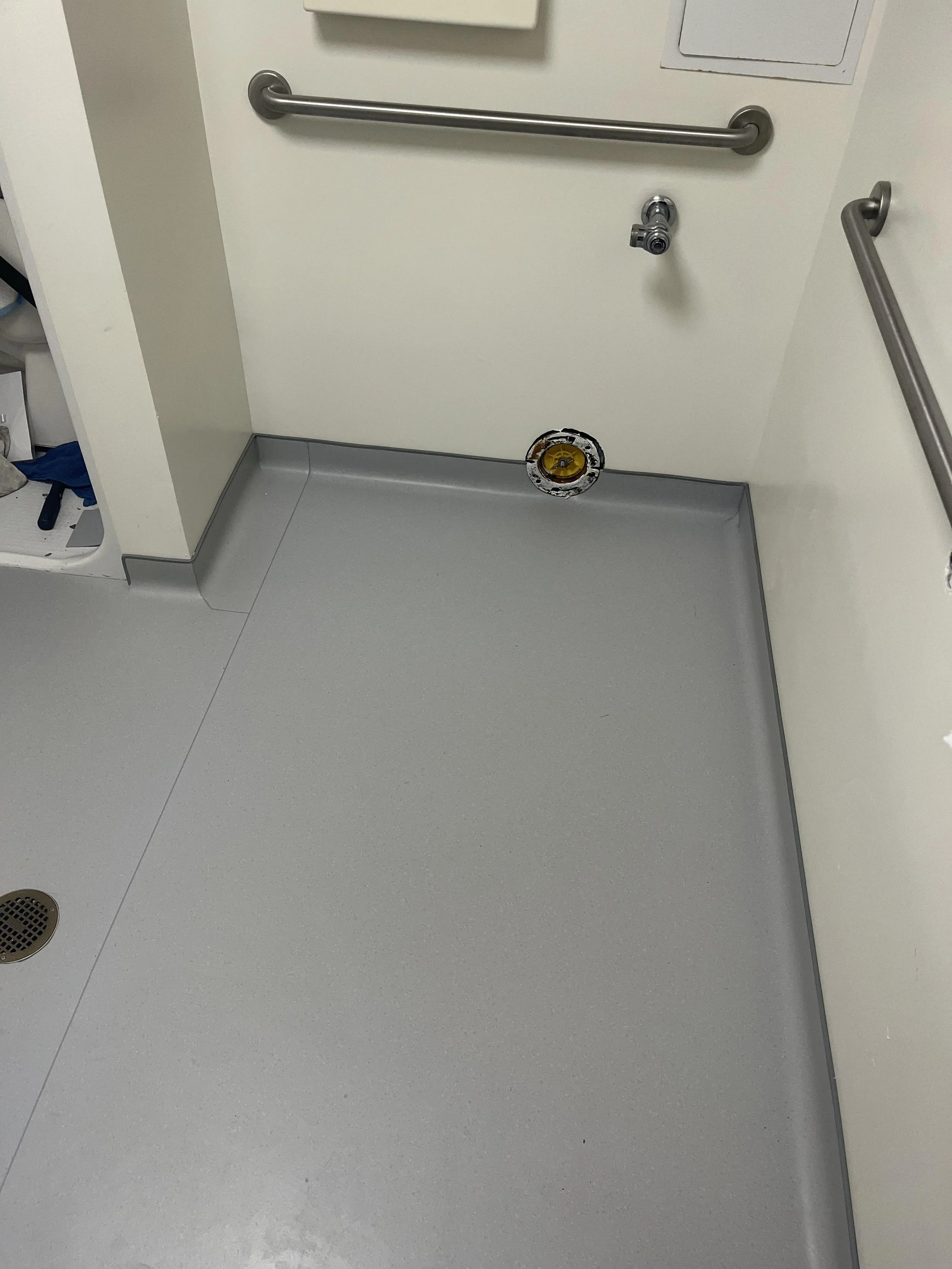 Hospital room floor with no mattress on the bed frame, a drain on the floor, and handrails on the wall.