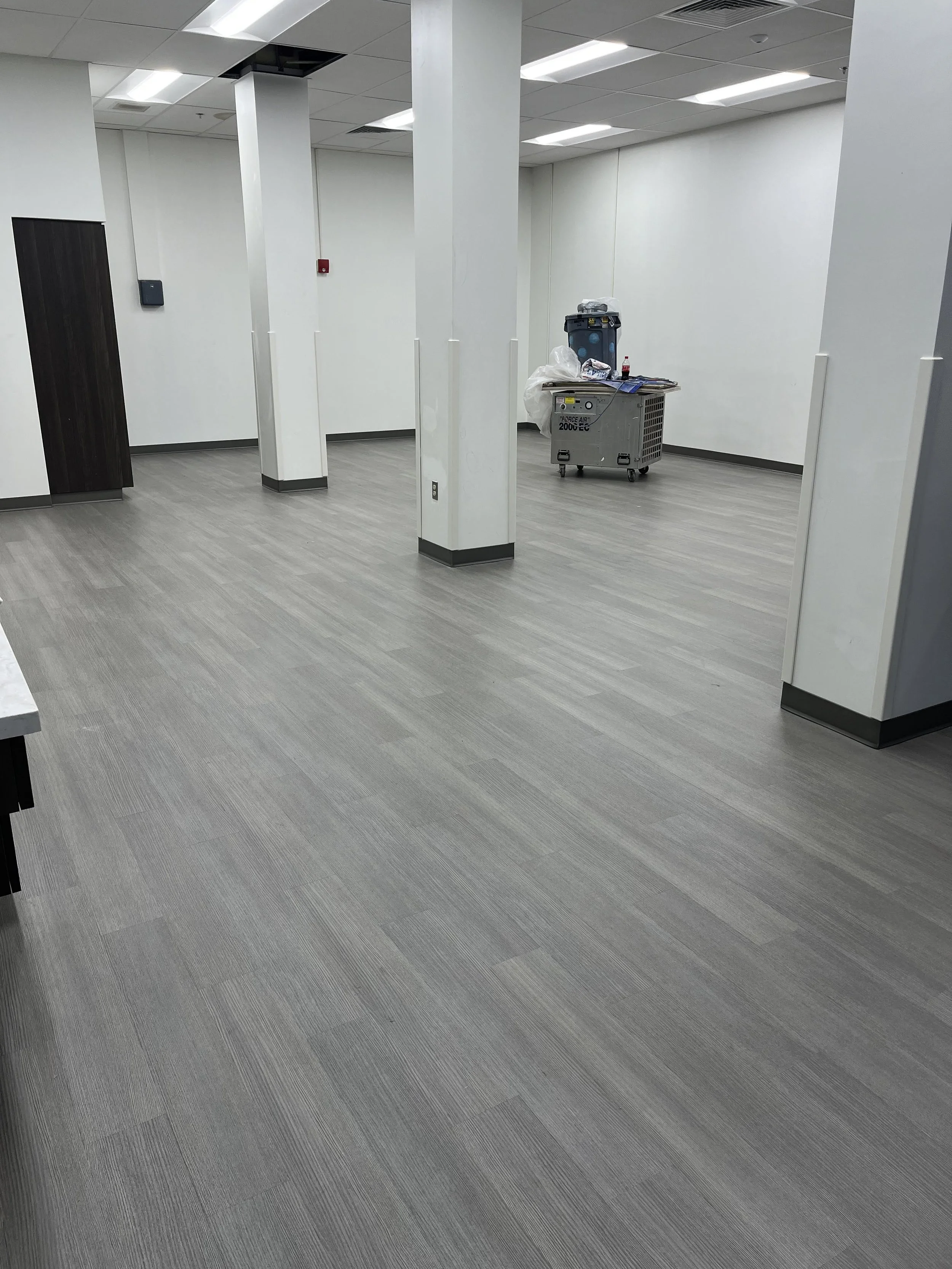 Empty office or commercial space with white walls, ceiling tiles, gray flooring, and a couple of columns. There is a cart with cleaning supplies and some trash bags in the back corner.