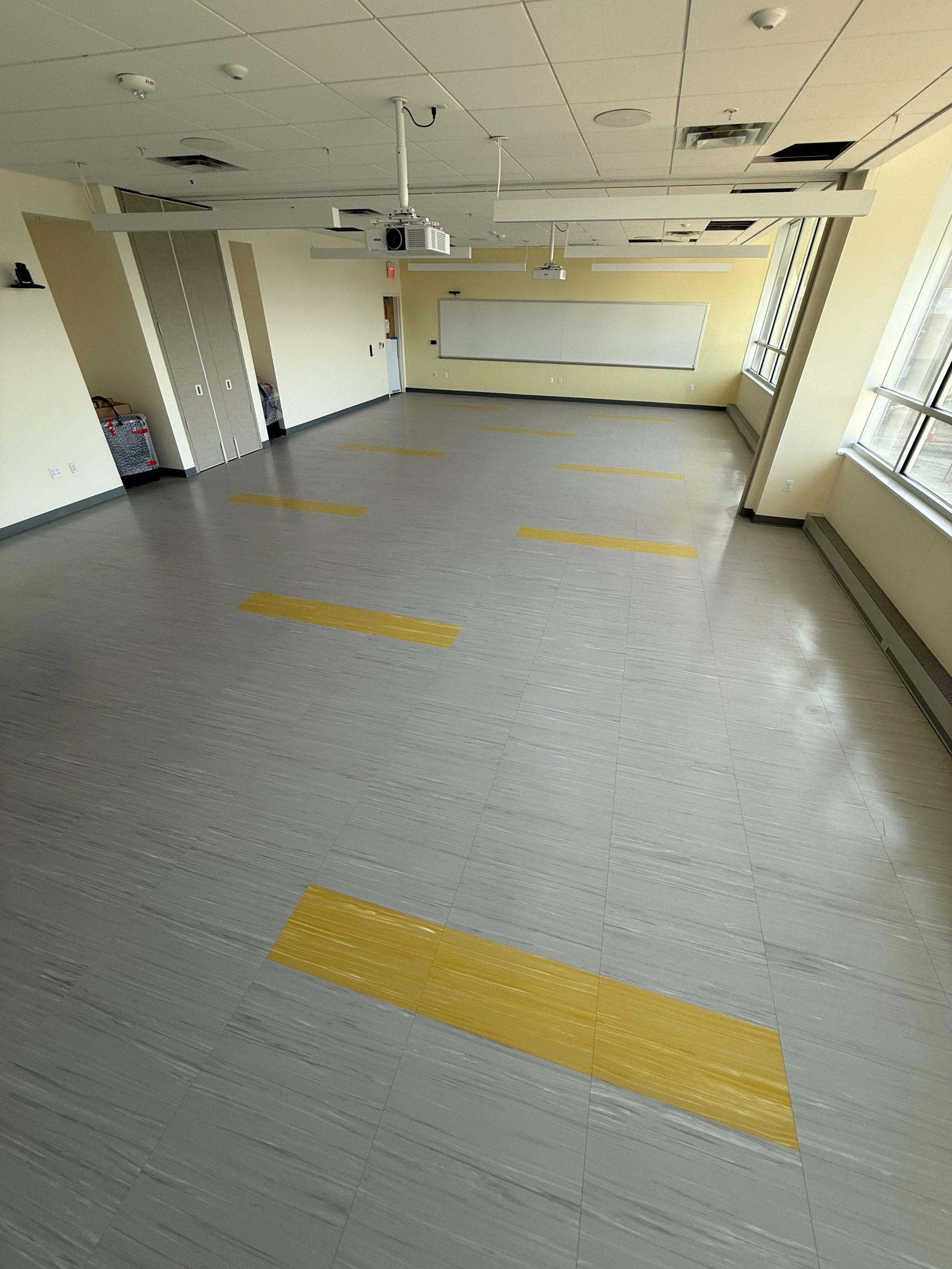 Empty classroom with yellow tactile paving strips on the gray floor, large windows on the right side, whiteboard at the front, and ceiling-mounted projectors.