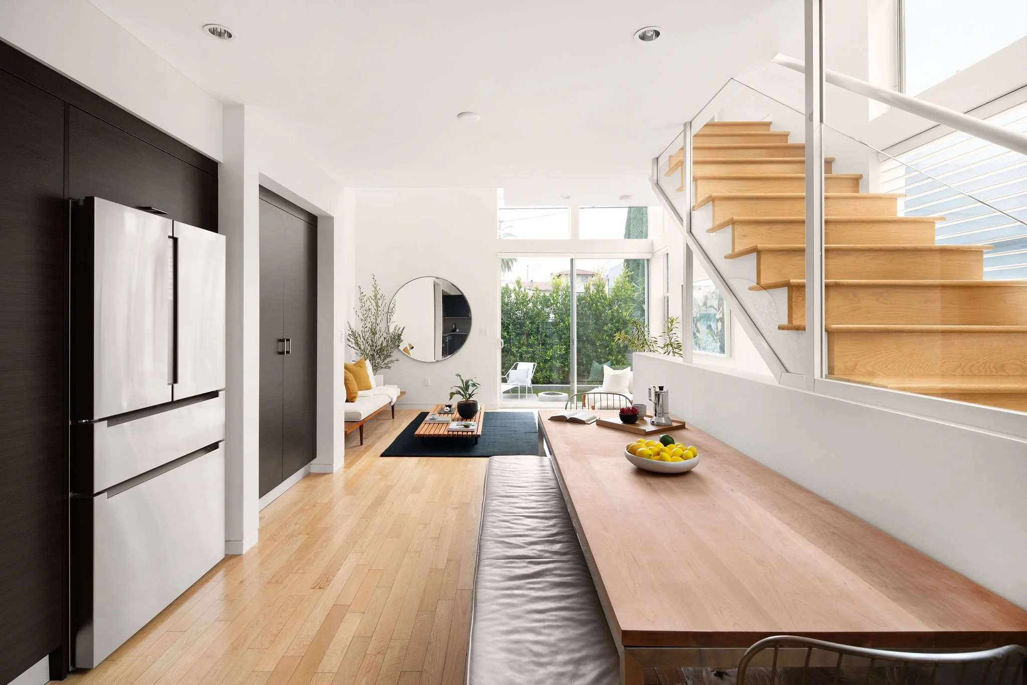 A bright modern living room with wooden floors, a staircase, and large windows overlooking greenery.