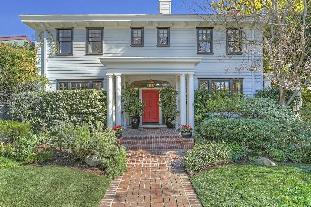 Front of a two-story house with white exterior and dark window frames, a red front door, and a brick walkway leading to the porch, surrounded by greenery and trees.