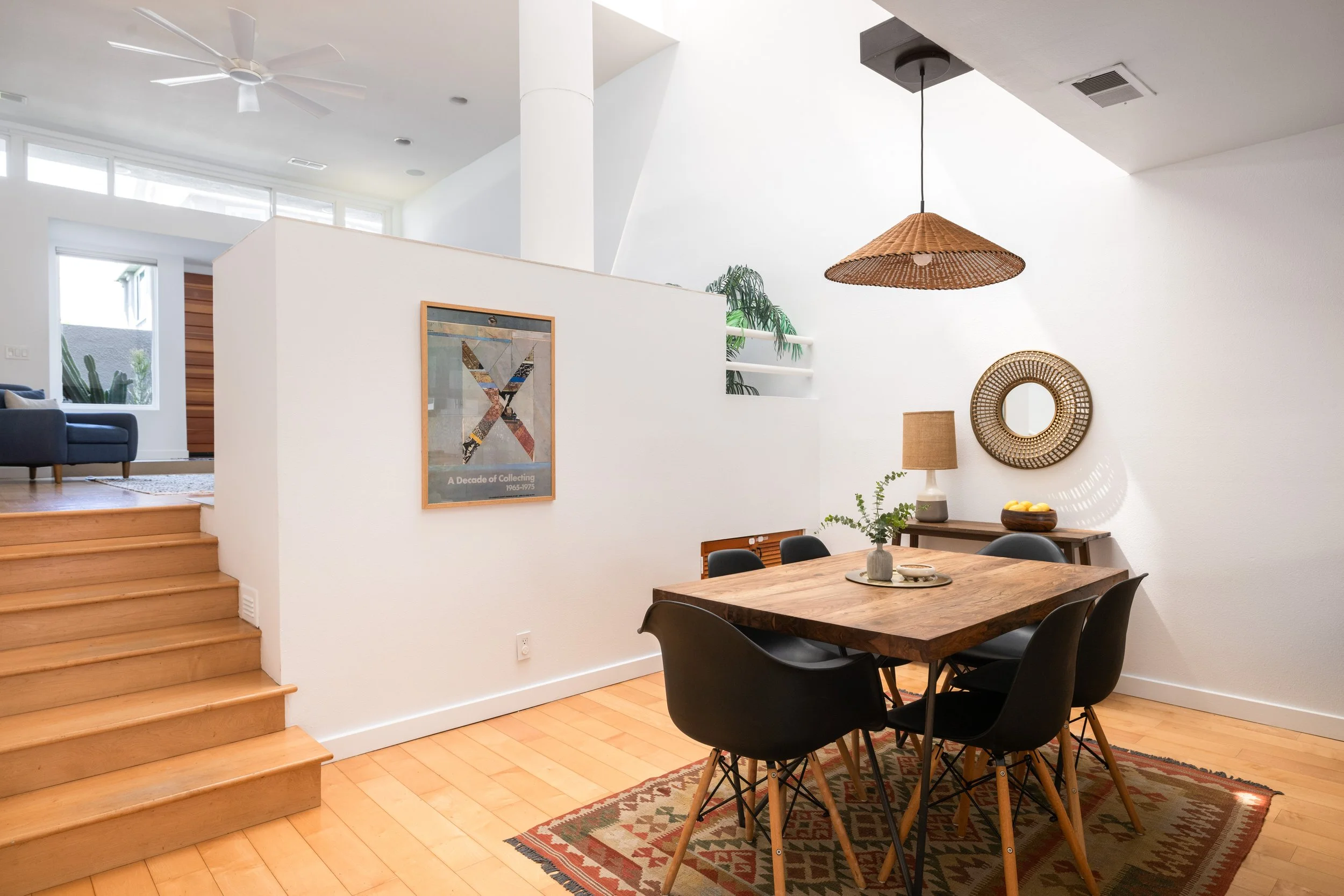 A dining room with a wooden table and six black chairs, a patterned rug, a sideboard with a lamp and a bowl of lemons, a round mirror, a rattan pendant light, and a staircase leading to a living area with a blue sofa and large windows.