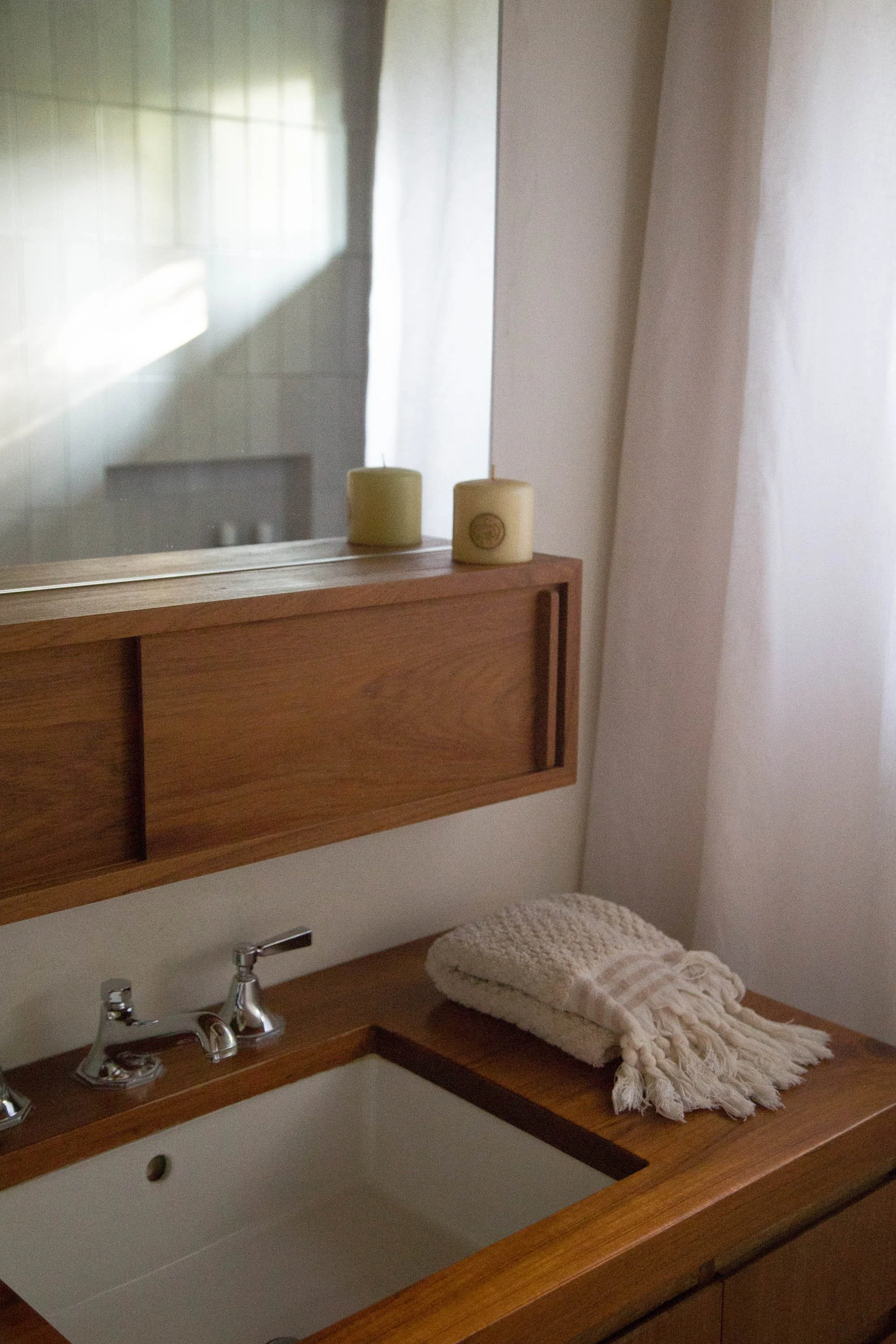 Bathroom sink with folded towels on wooden countertop, mirror with two candles and white curtains in the background.