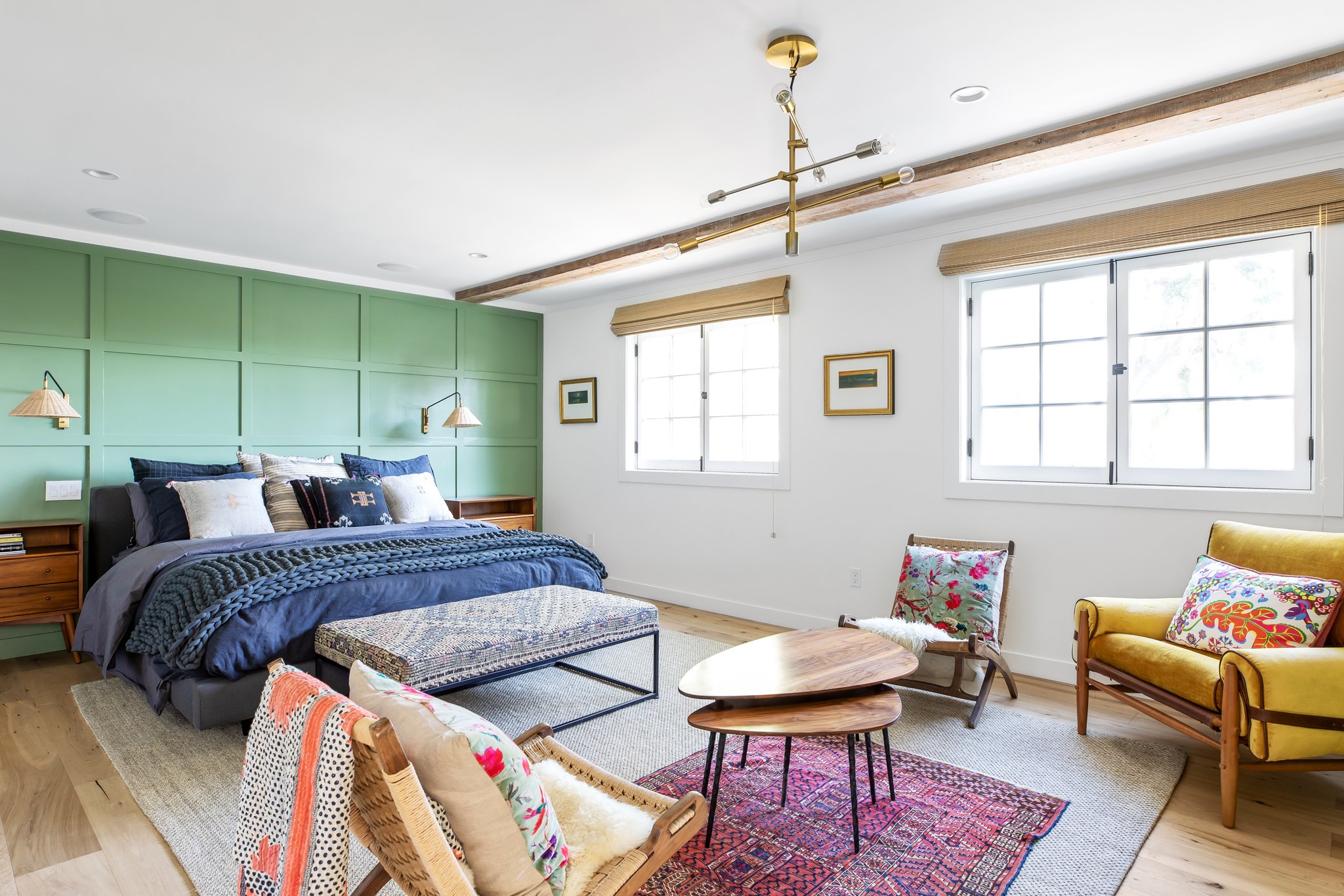 A bedroom with a green paneled wall behind a bed with multiple pillows and a navy blue comforter. There are two wooden nightstands, wall-mounted lamps, and two windows with beige shades. The room has colorful chairs, a wooden coffee table, a woven be