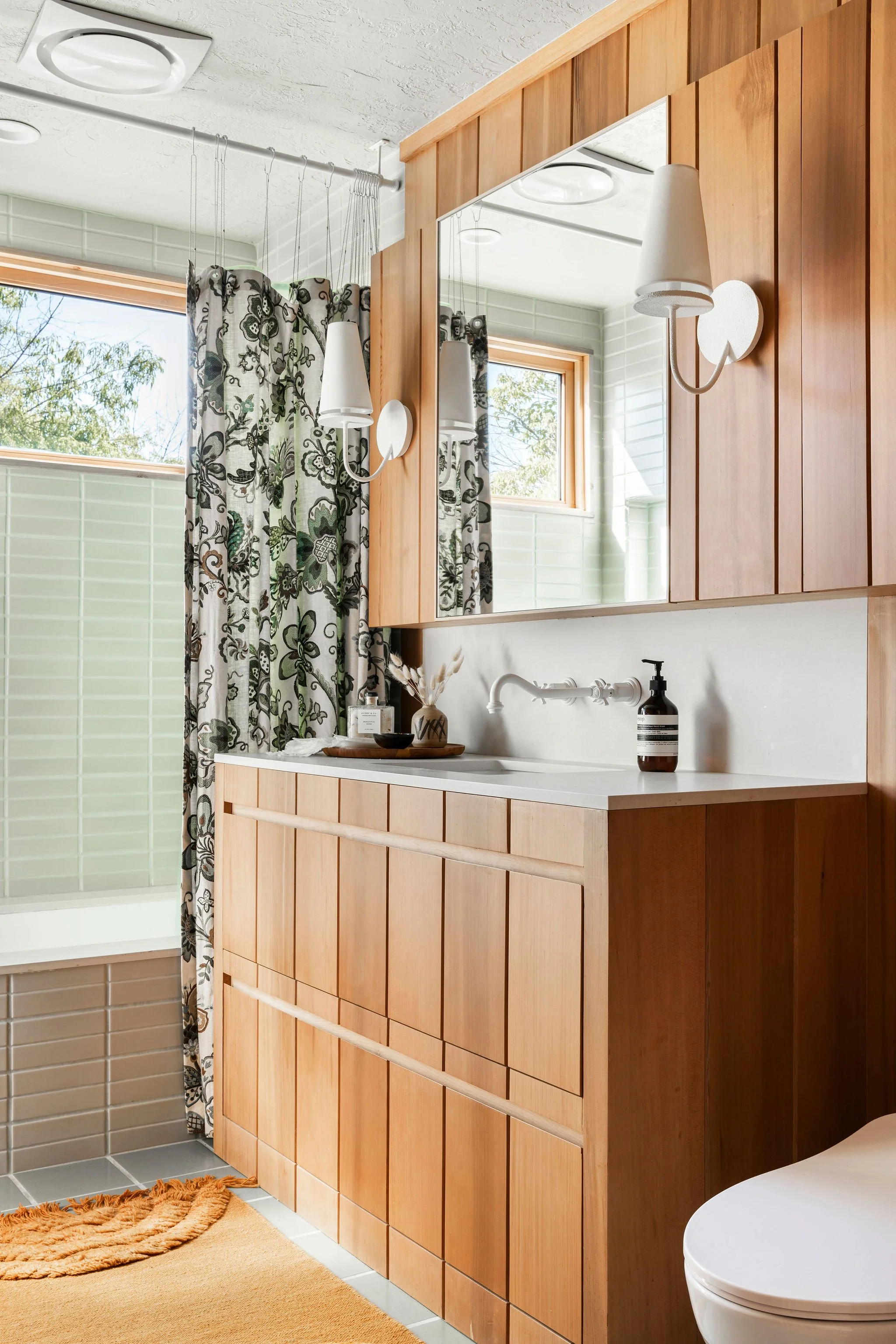 A modern bathroom with wooden cabinetry, a white countertop, a mirror, a window with a curtain, a wall-mounted light, and a small section of a white toilet. There is a soap dispenser on the counter and a small vase with dried flowers.