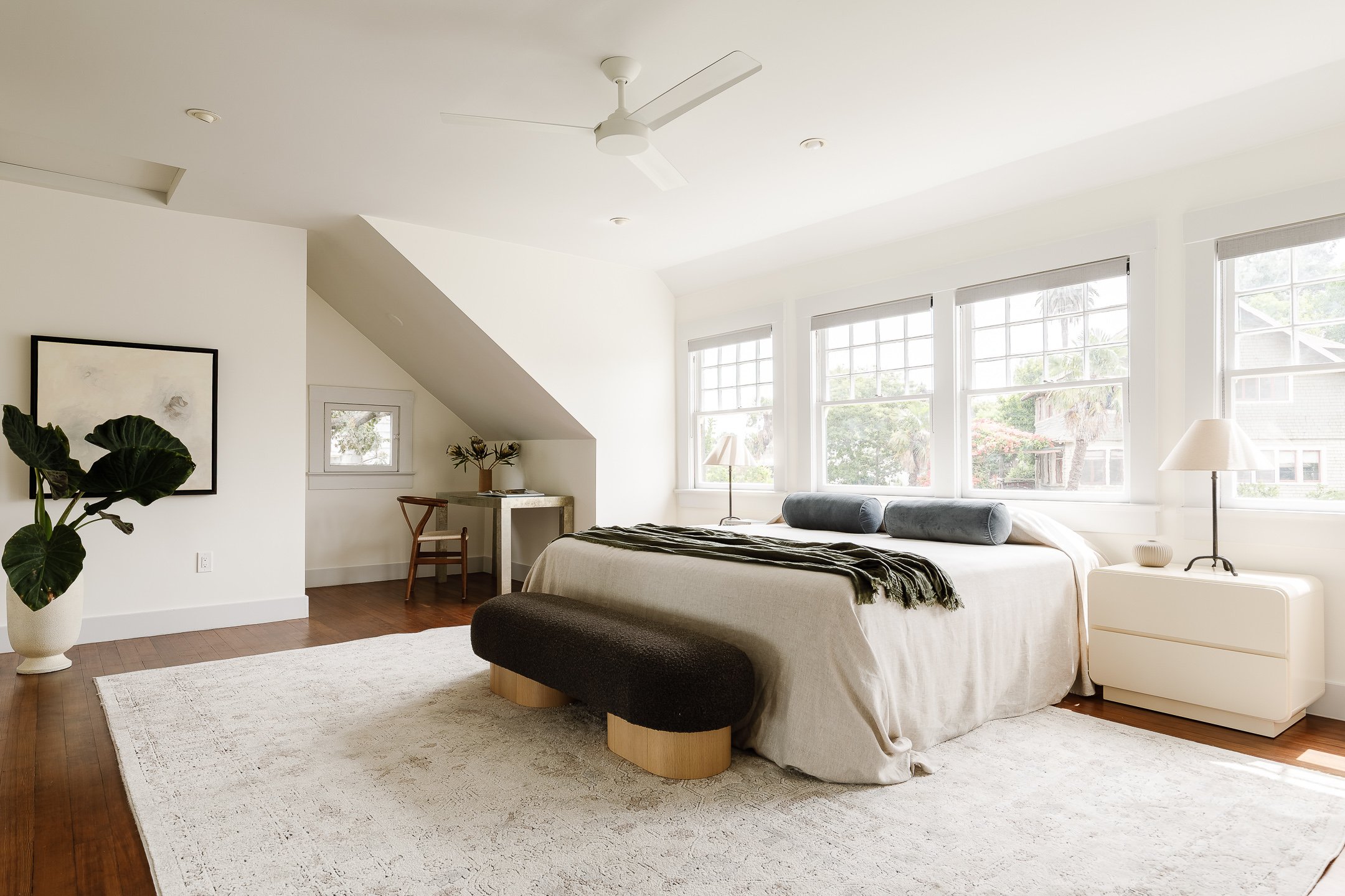 Bright bedroom with large windows, white walls, a bed with white bedding, gray pillows, and a dark bench at the foot of the bed. A white nightstand with a lamp and small decor, a potted plant, a framed artwork, and a small desk with a chair are also 