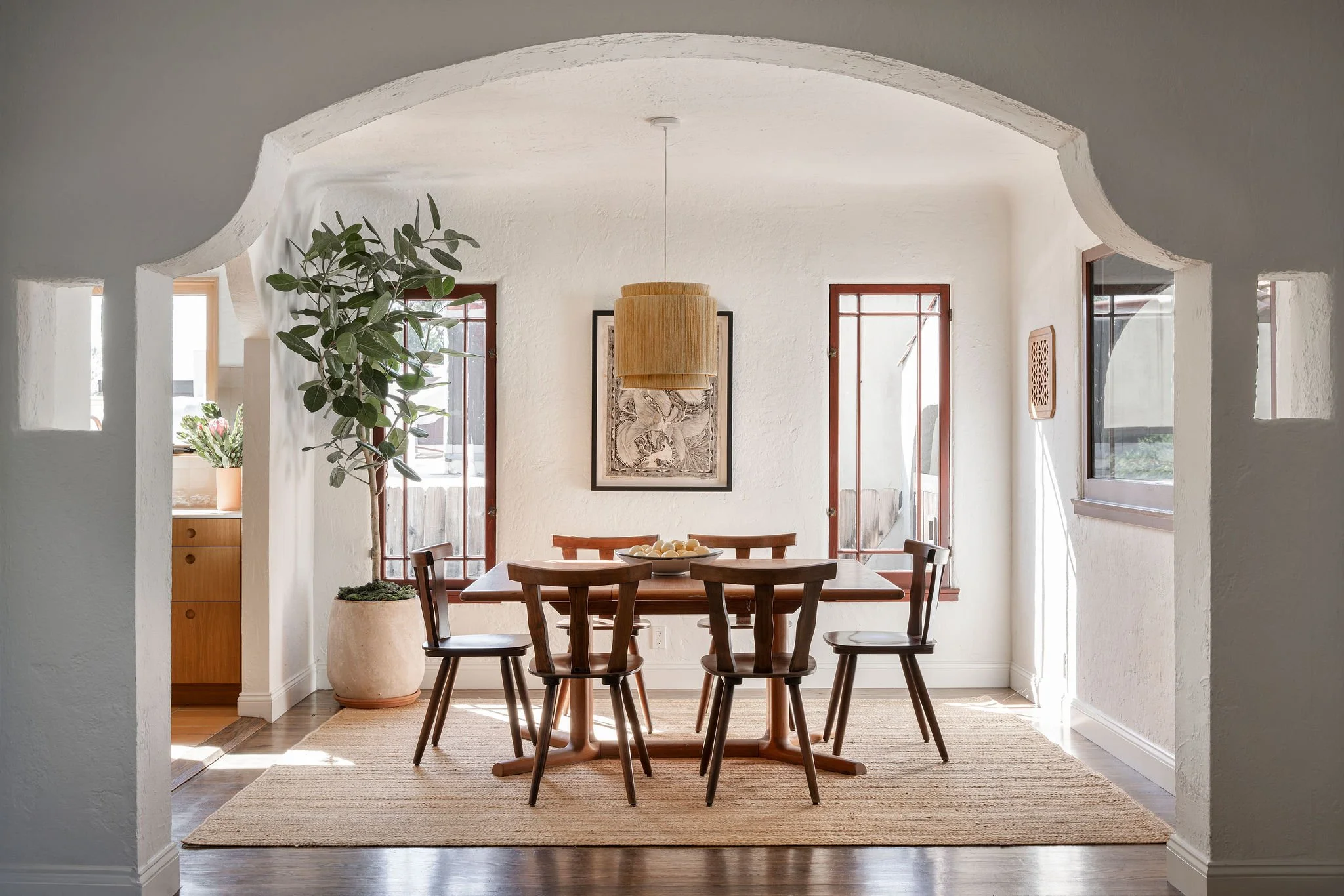 A dining room with a wooden table and six matching chairs, a potted plant, framed artwork, and natural light from windows.