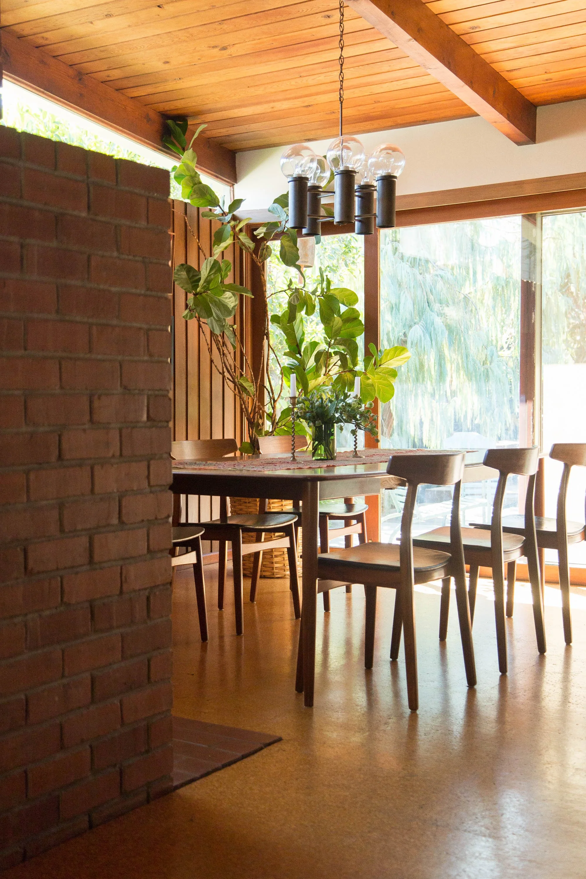 Bright dining area with wooden ceiling and furniture, large windows with natural light, potted plants on the table, and a modern chandelier hanging from the ceiling.