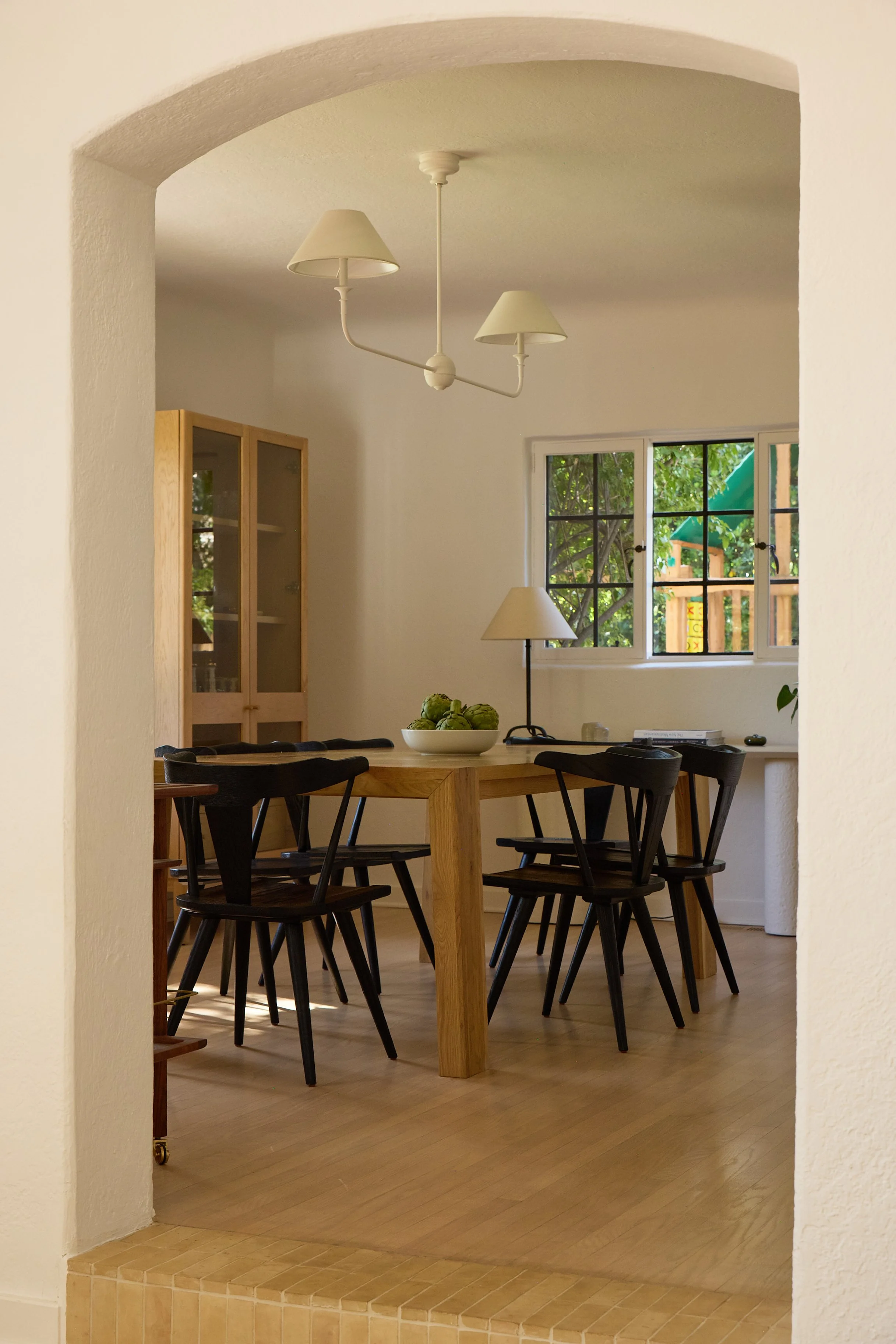 Dining room with wooden table, six black chairs, a bowl of artichokes, and a window showing greenery outside.