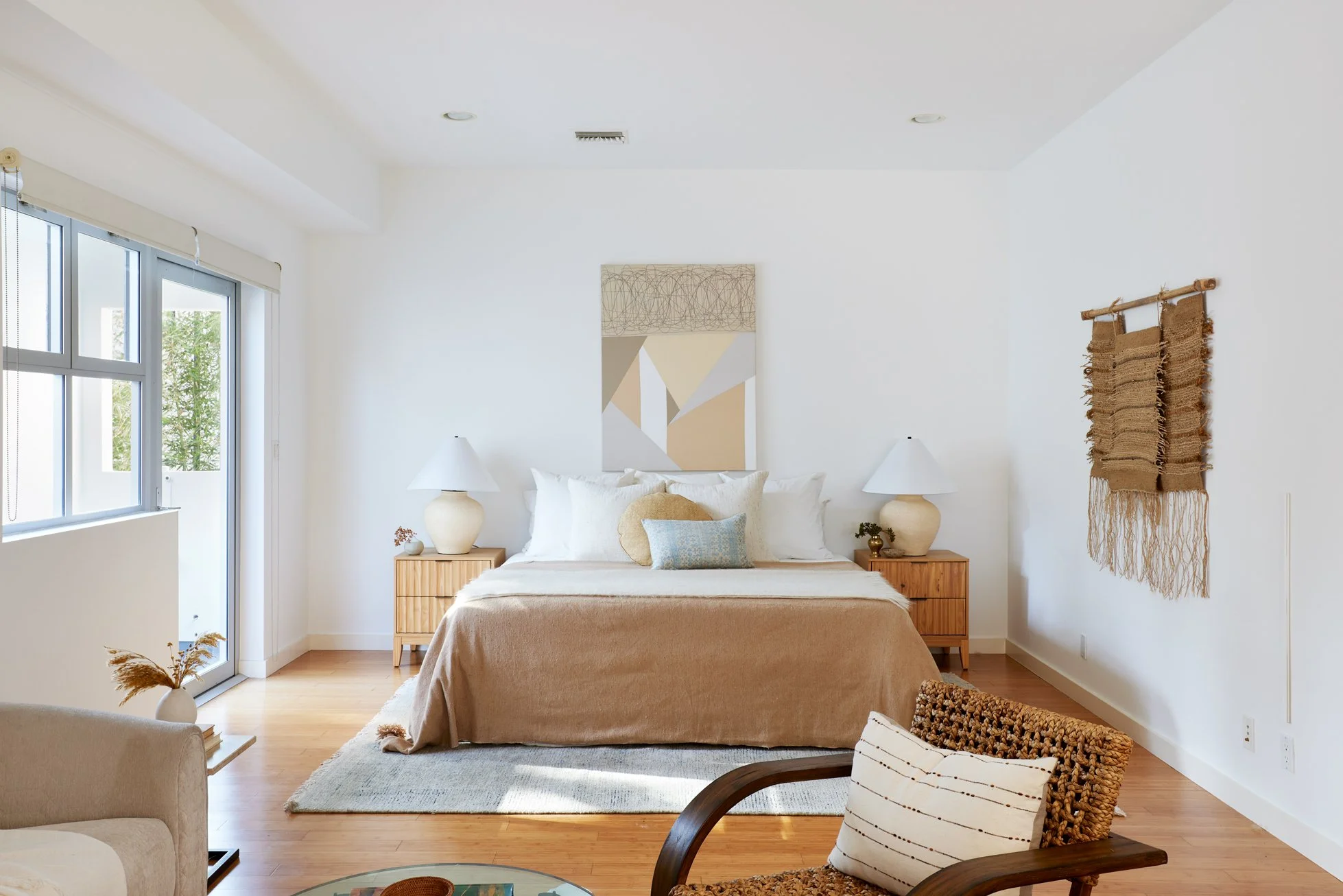 A bedroom with a large bed, wooden nightstands, and lamps on each side. There is abstract artwork above the bed, a window with blinds, and woven wall hangings on the wall. The room has a wooden floor and natural decor.