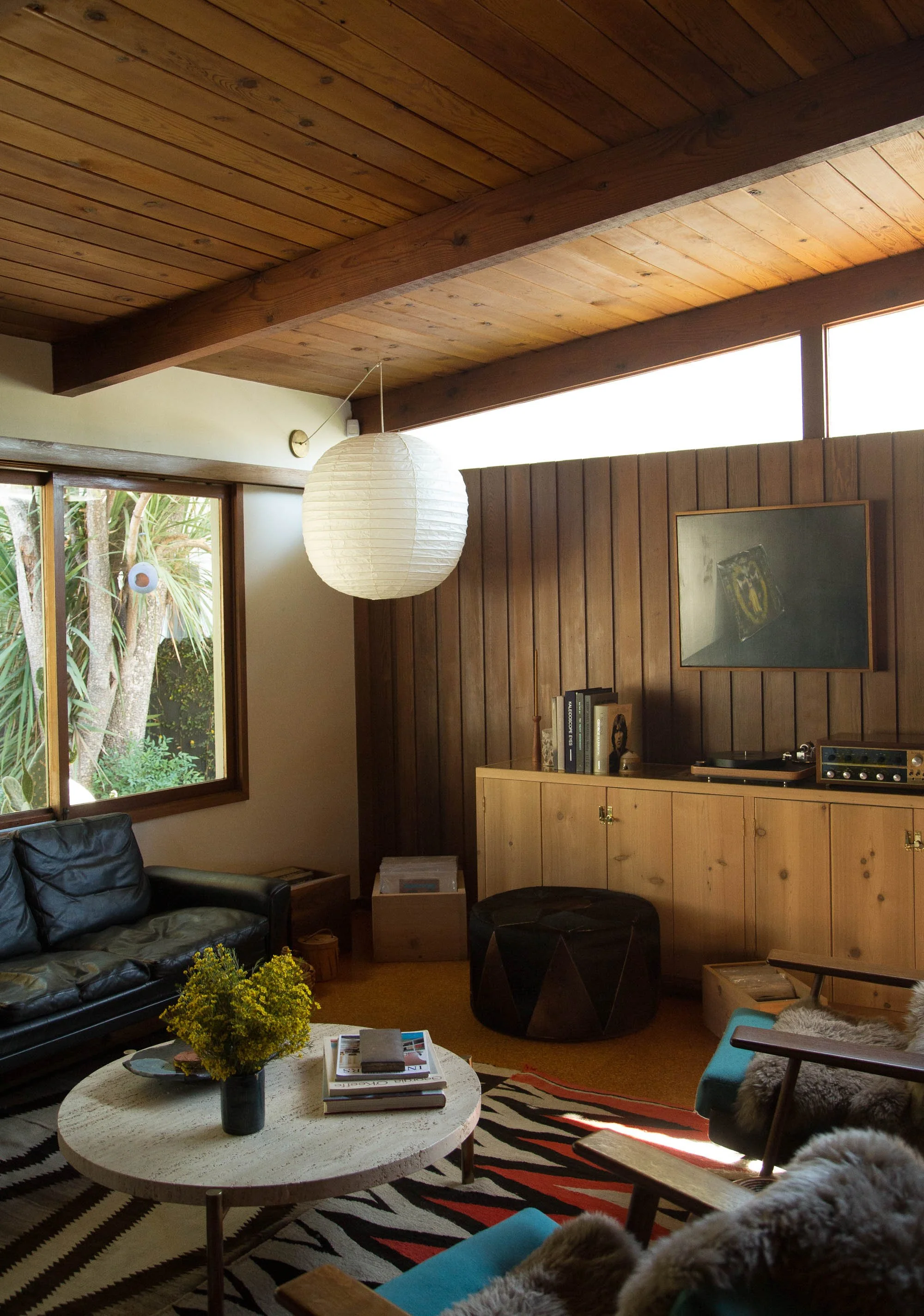 Living room with wooden ceiling and wall paneling, black leather sofa, armchair with sheepskin throw, round coffee table with magazines and yellow flowers, record player and vinyl on cabinet, window views of trees, paper lantern hanging