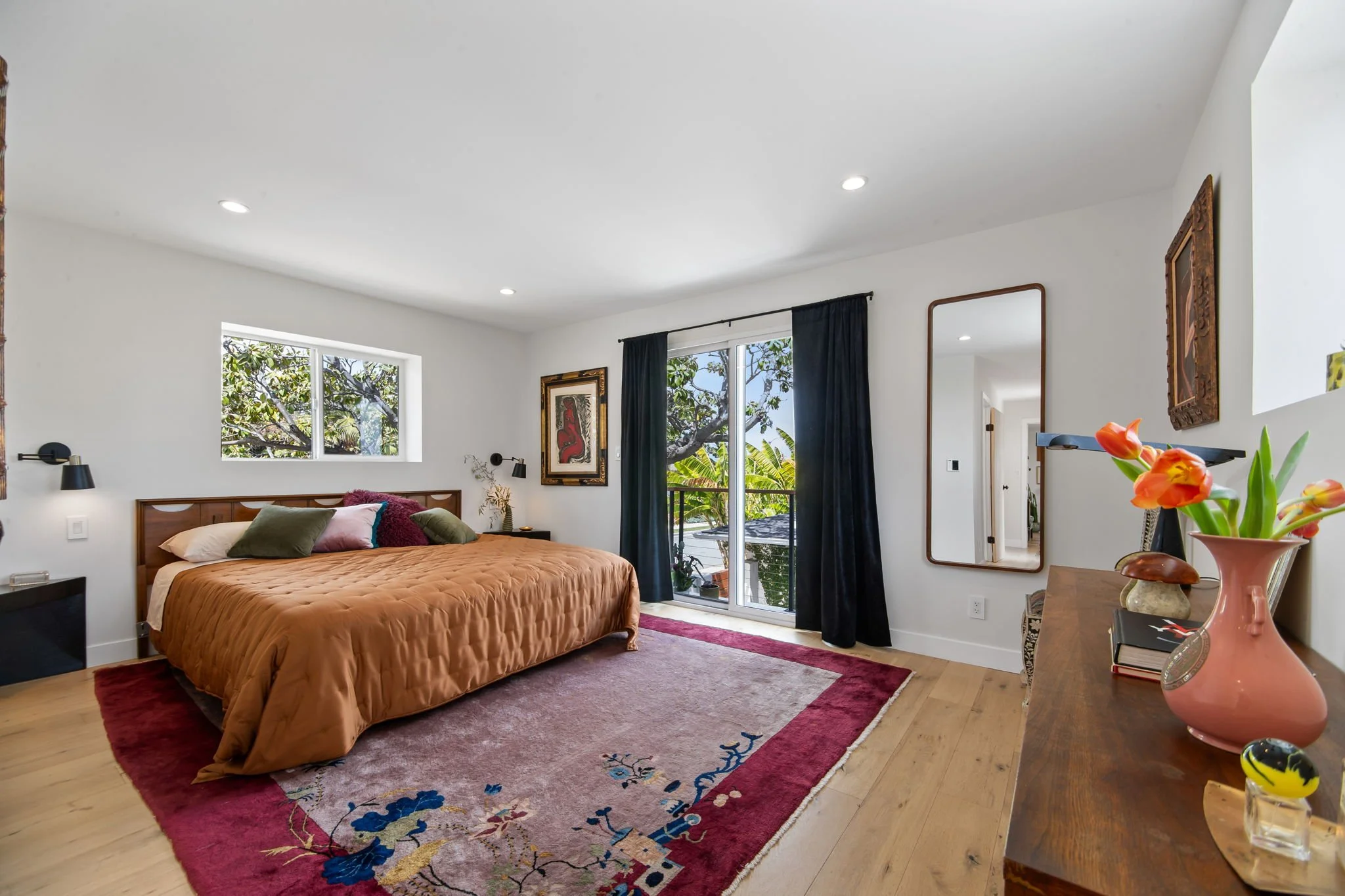 A bedroom with a large bed covered in brown bedding, multi-colored pillows, a pink rug with blue and gold accents, a window with trees outside, black curtains, and a wooden dresser with a pink vase and flowers.
