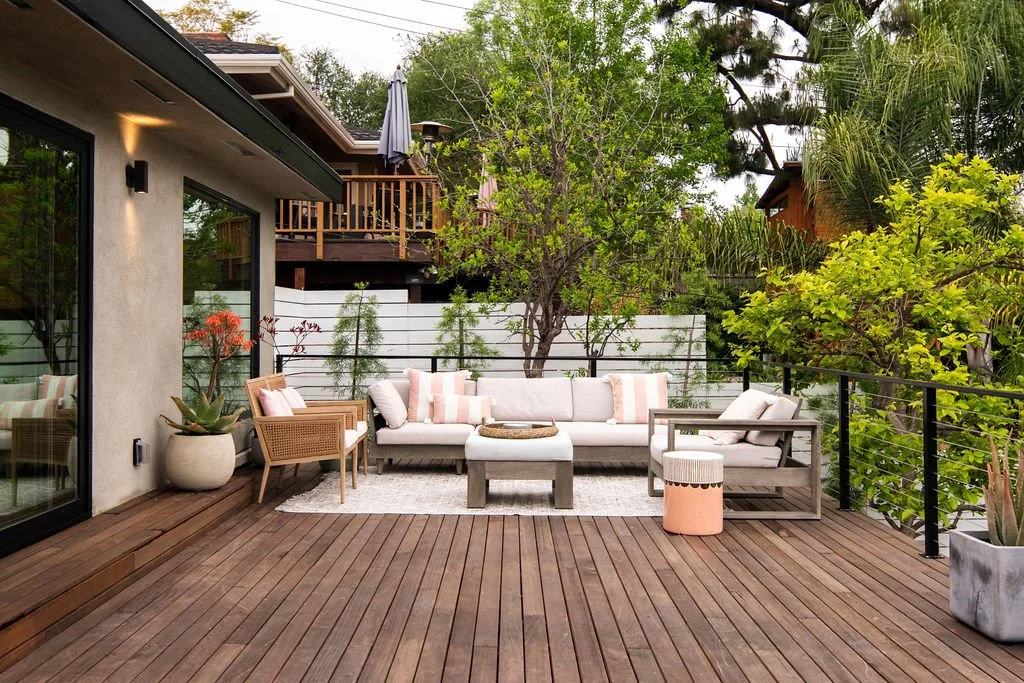 Outdoor wooden deck with beige outdoor furniture, cushions, potted plants, and surrounded by lush green trees and a white fence.