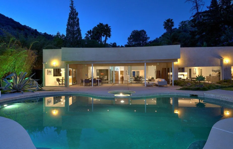 Modern house with large windows and outdoor seating area, illuminated at night, with a swimming pool in the foreground and trees in the background.