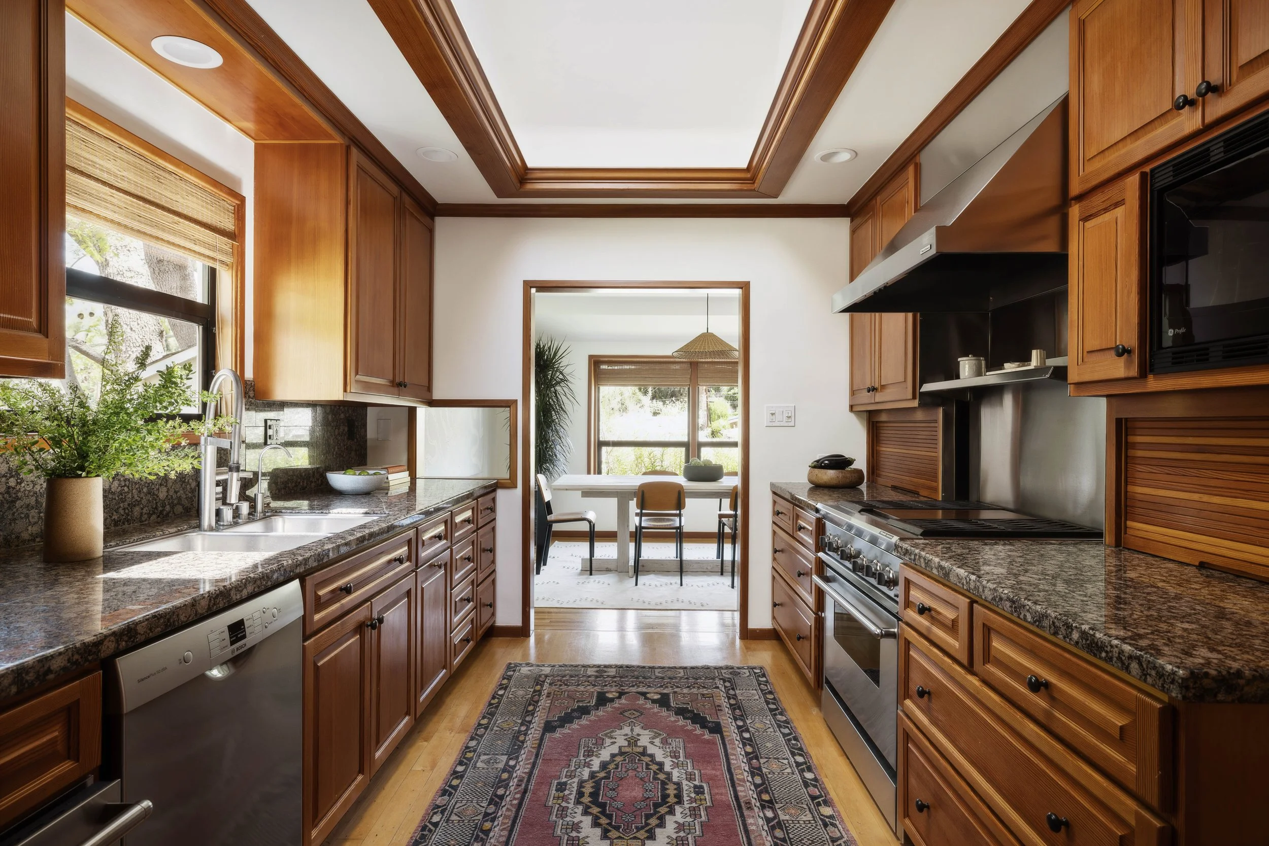 A kitchen with wooden cabinets, granite countertops, stainless steel appliances, and a window with a view of the outdoors. There is a dining area with a table and chairs visible through the doorway.
