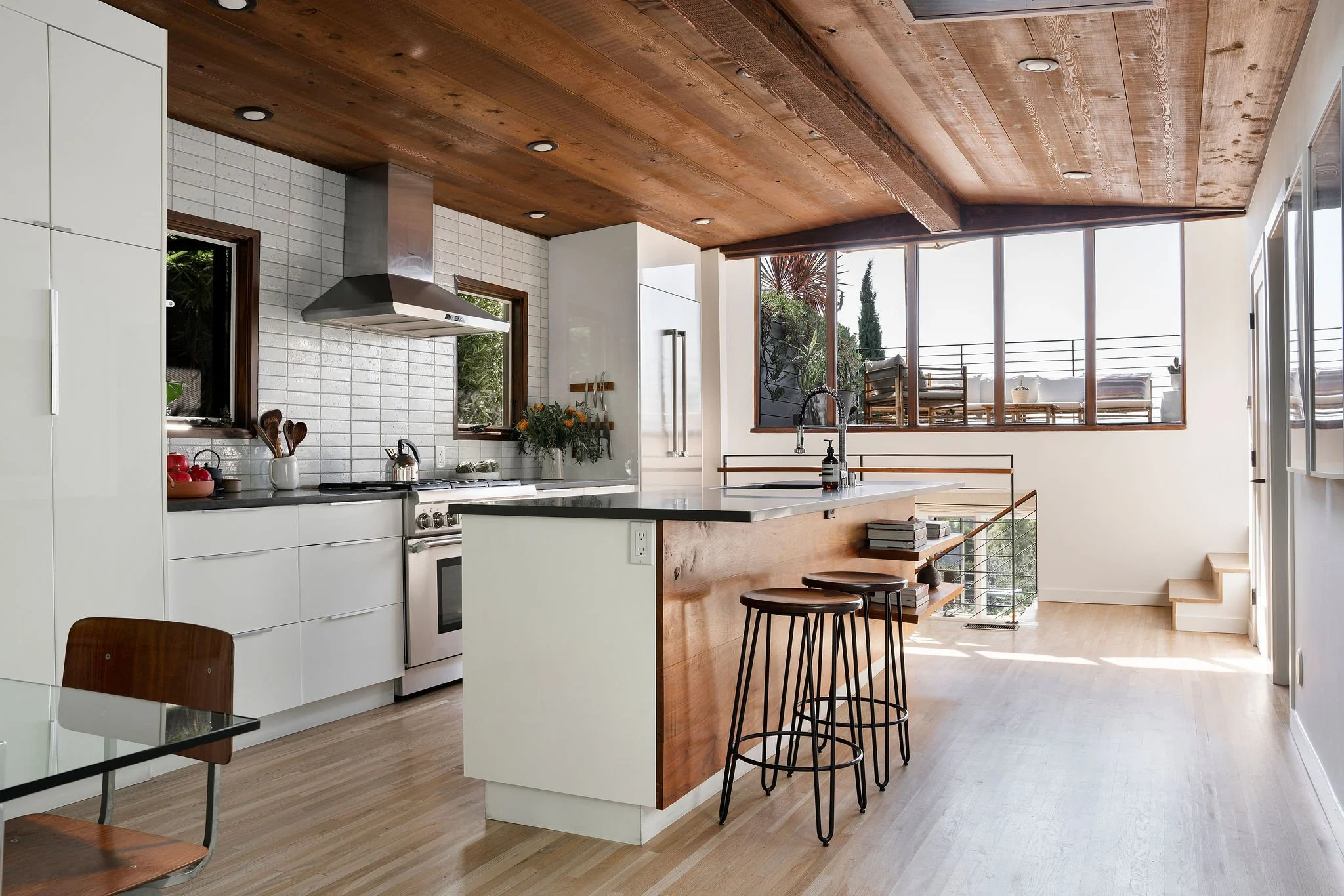 Modern kitchen with white cabinets, wooden ceiling, and large windows. Includes a kitchen island with bar stools and a view of the outdoors.