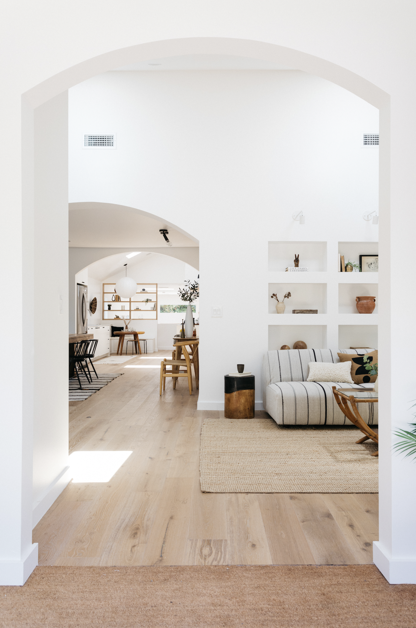 Living room with white walls, built-in shelves, striped sofa, wooden coffee table, and beige rug, leading into a dining area with wooden chairs and hanging light fixture, all in a minimalist style.