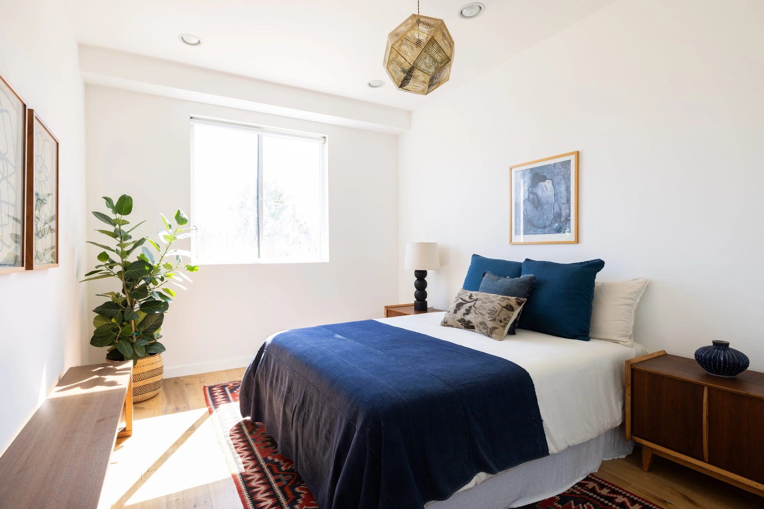 Bright bedroom with a large window, white walls, and wooden furniture. The bed has white sheets, a navy blue blanket, and multiple decorative pillows. There is a tall green potted plant on the left, a wooden nightstand with a black lamp and a framed 