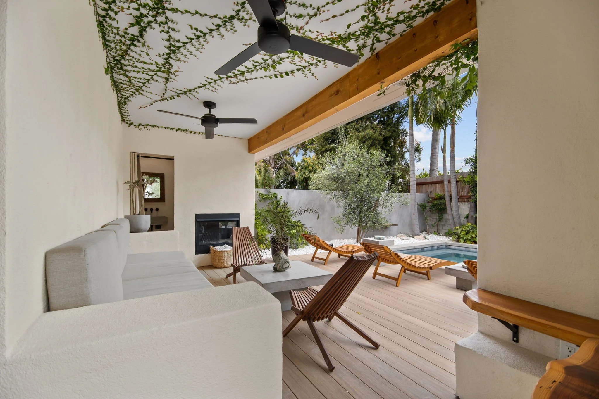 Outdoor patio with a white couch, wooden chairs, a rectangular table, a pool, and lush green plants, including palm trees, under a ceiling with hanging vines and ceiling fans.