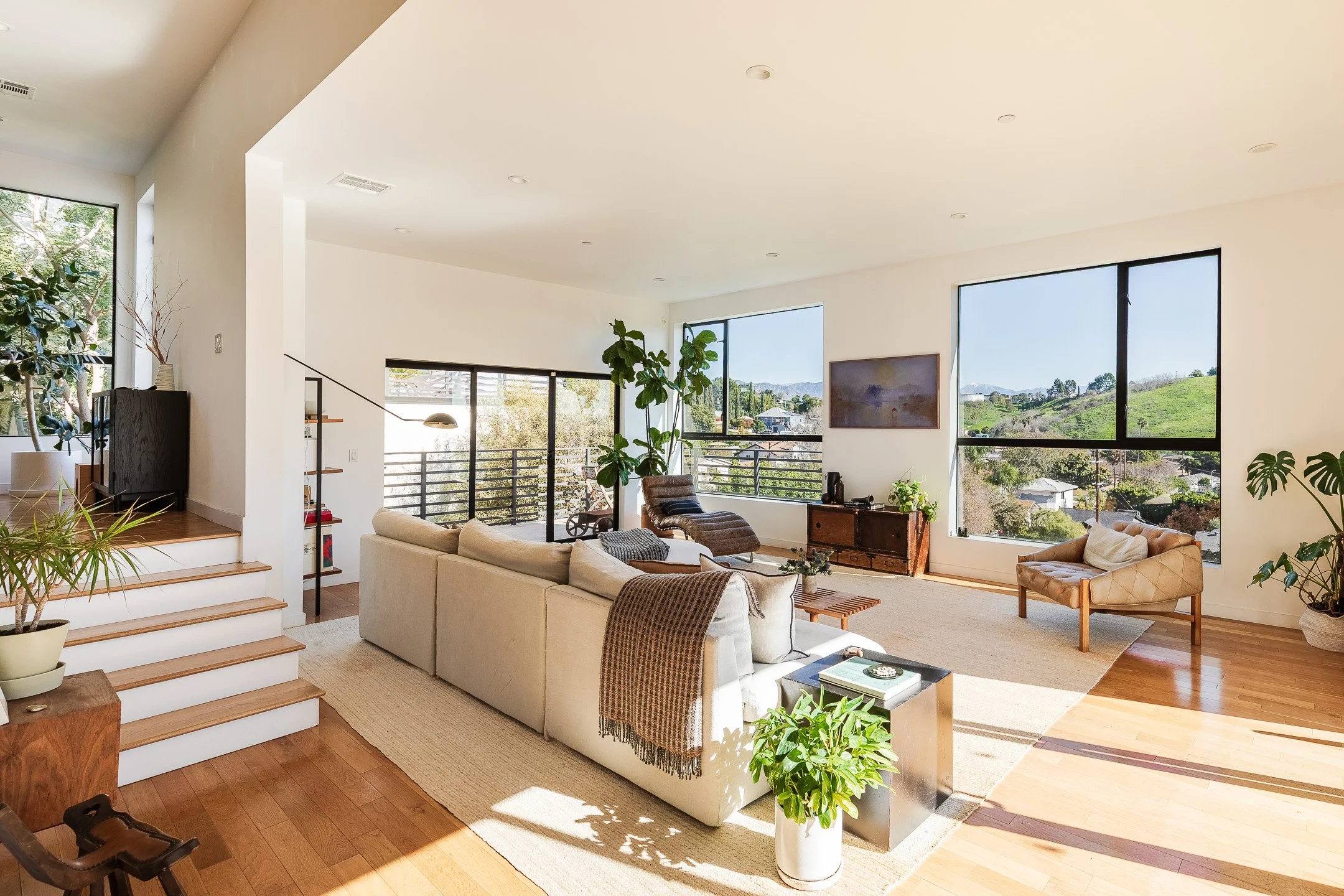 Bright living room with large windows showing a scenic hillside view, modern beige sofa, wooden floors, and various houseplants.