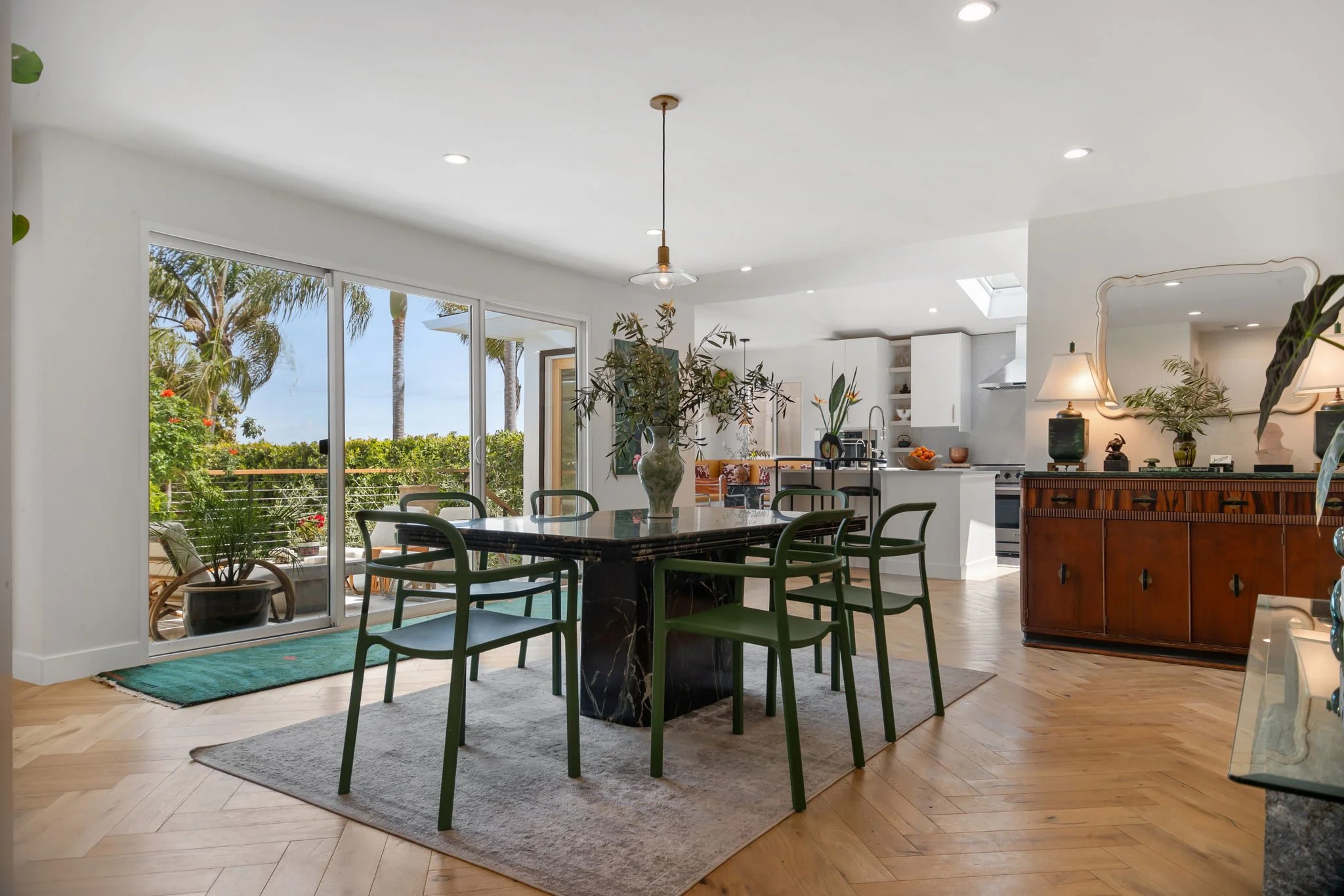 Bright open-concept dining area with a large table, green chairs, a vase with flowers, and sliding glass doors leading to a patio with outdoor furniture and palm trees.