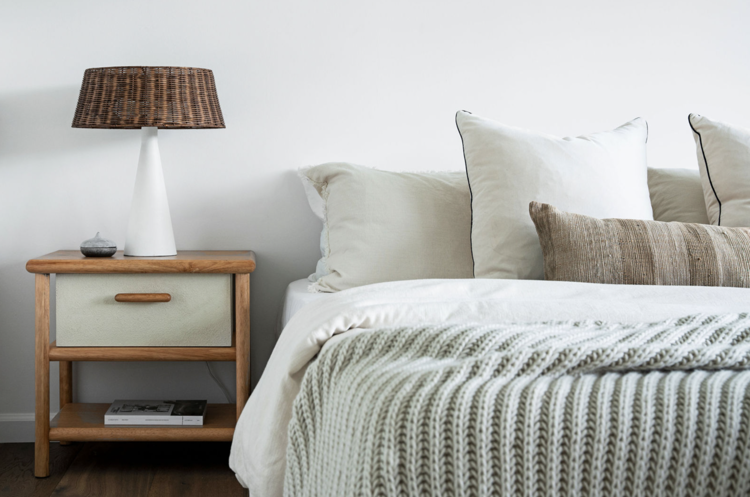 A bedroom with a white bed, multiple pillows, a knitted blanket, a wooden nightstand with a drawer and a shelf, a white lamp with a woven brown lampshade, and a small decorative stone on the nightstand.