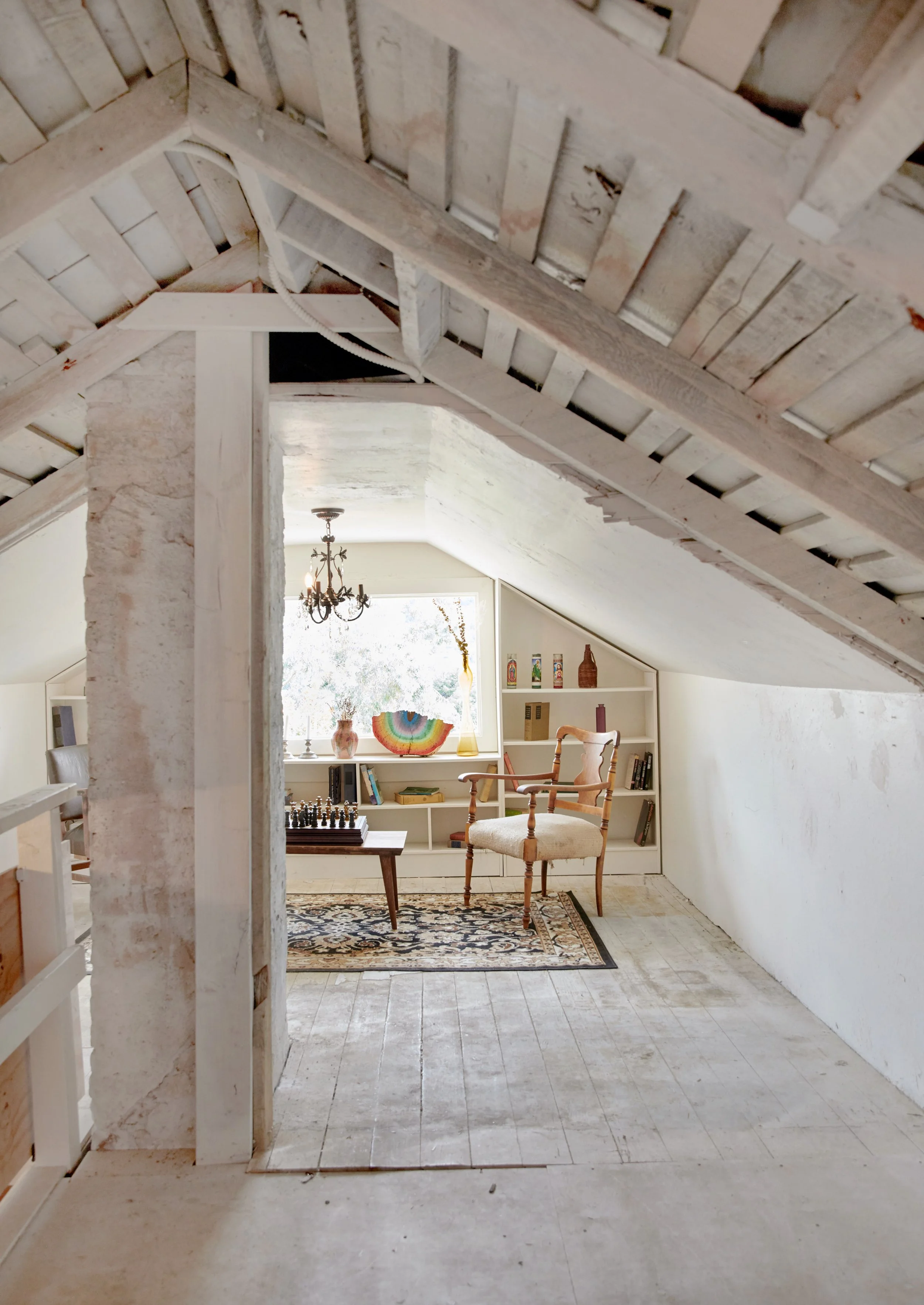 An attic room with slanted ceiling, white walls, and wooden flooring. There is a small area rug, a vintage wooden armchair with a cushion, a wooden table with a chessboard, and a built-in bookshelf with decorative items and books. A chandelier hangs 