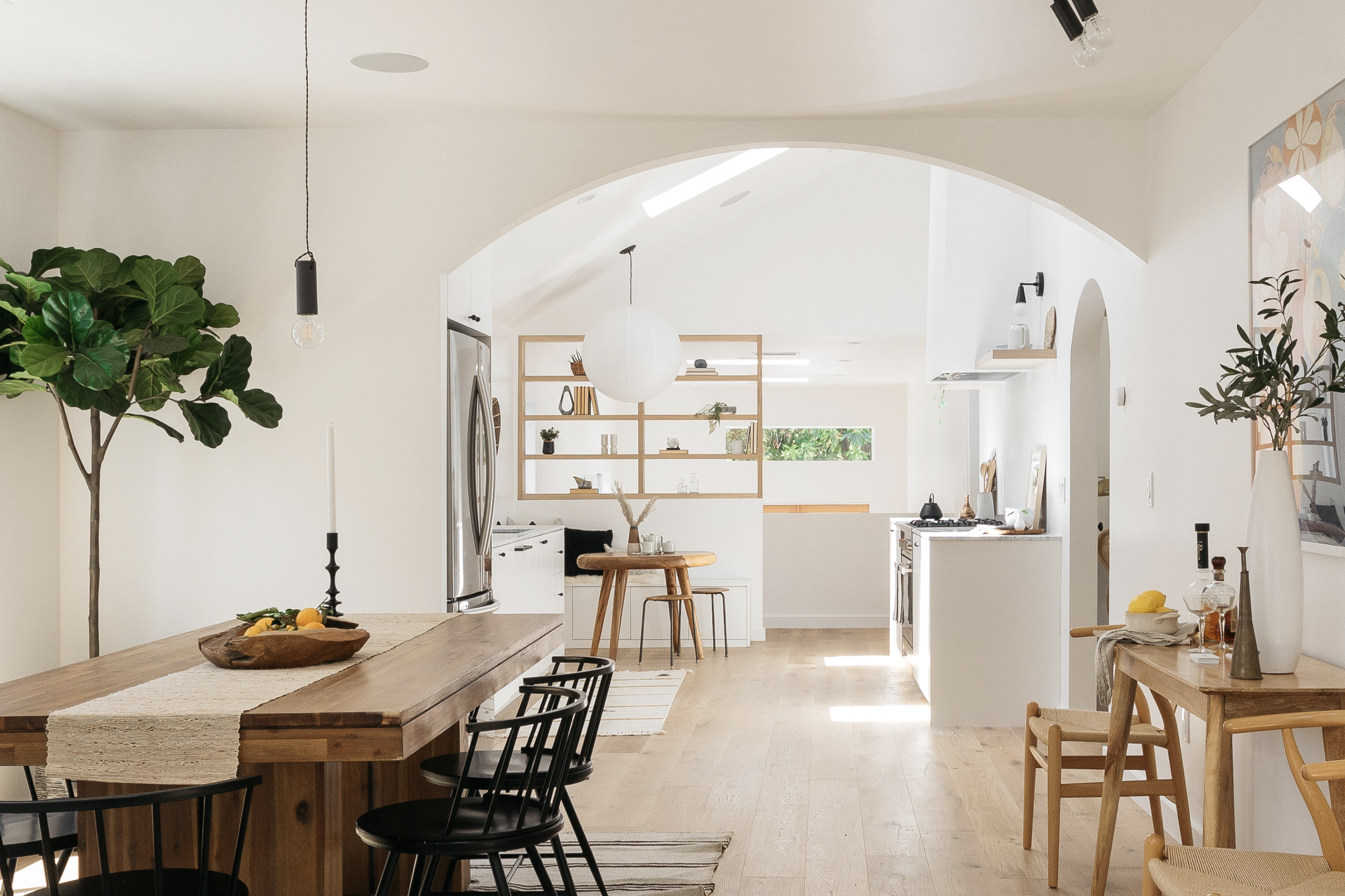 Bright, minimalist open-plan kitchen and dining area with light wood accents, a large wooden dining table with black chairs, a small round wooden table with a stool, decorative shelves, and green plants.