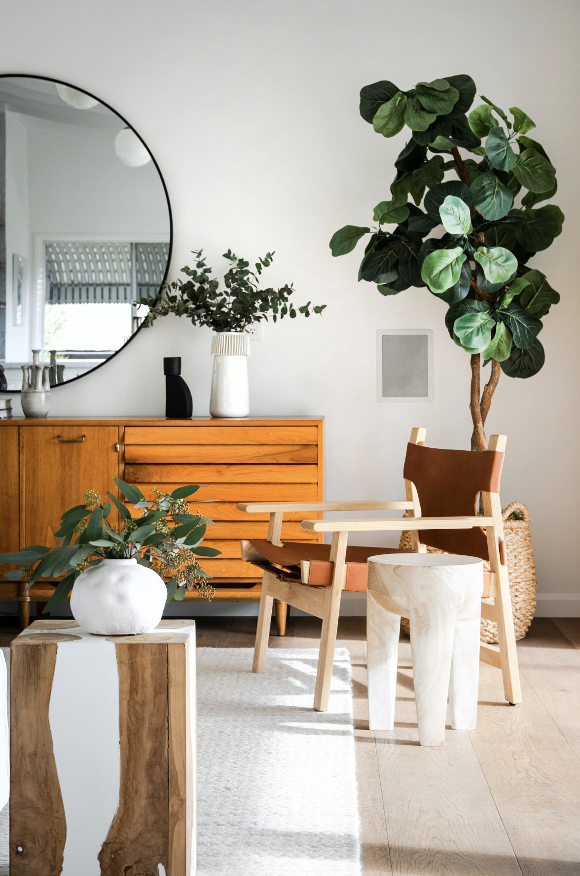 Interior of a modern living room with a round mirror, wooden sideboard, large potted plant, and decorative vases and furniture, featuring natural light and minimalist decor.