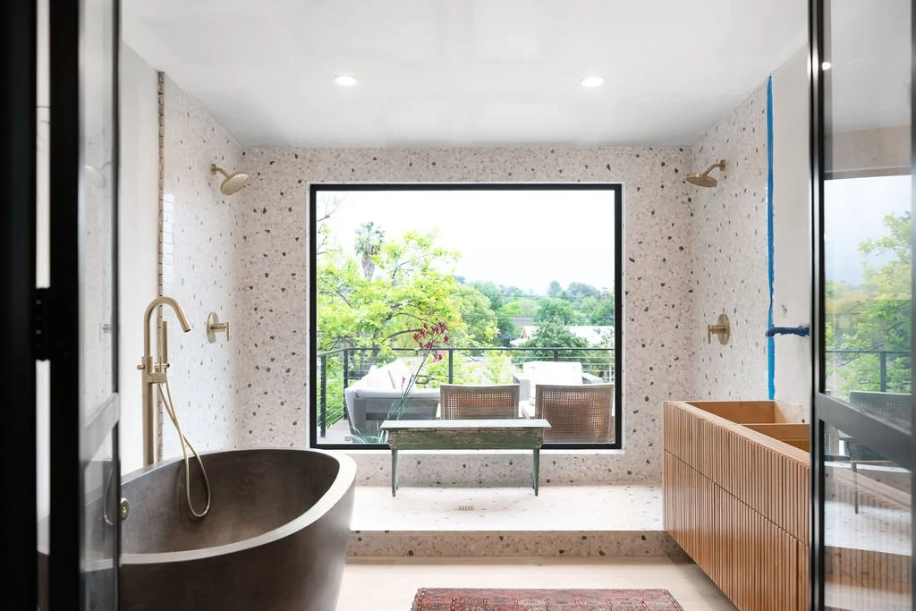 A modern bathroom with a round freestanding bathtub, a large window with a view of greenery, and an outdoor balcony with seating.