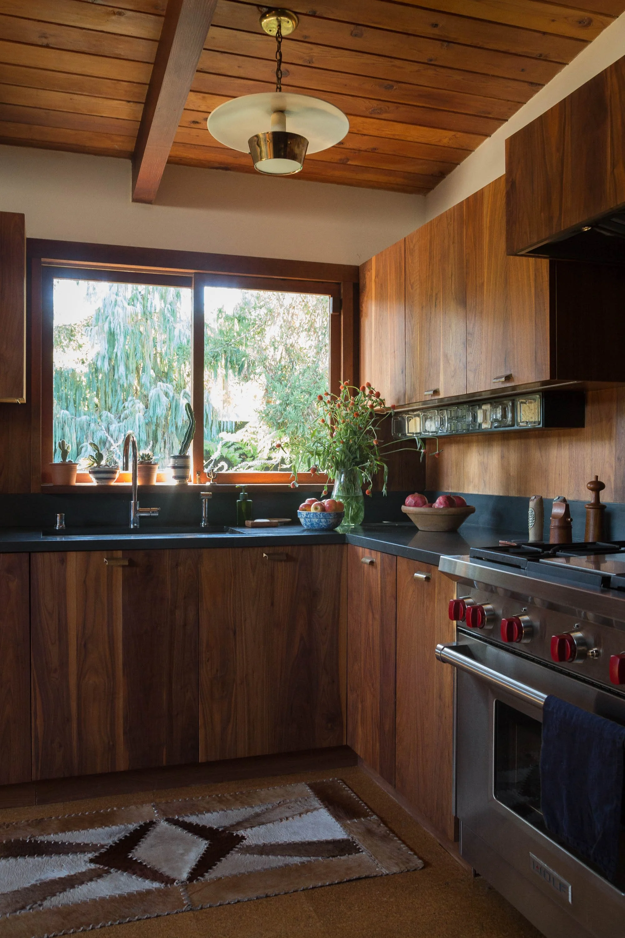 A cozy kitchen with wooden cabinets, a black countertop, a window with a view of greenery, potted plants, a vase with flowers, bowls of apples, and a stainless steel stove with red knobs.
