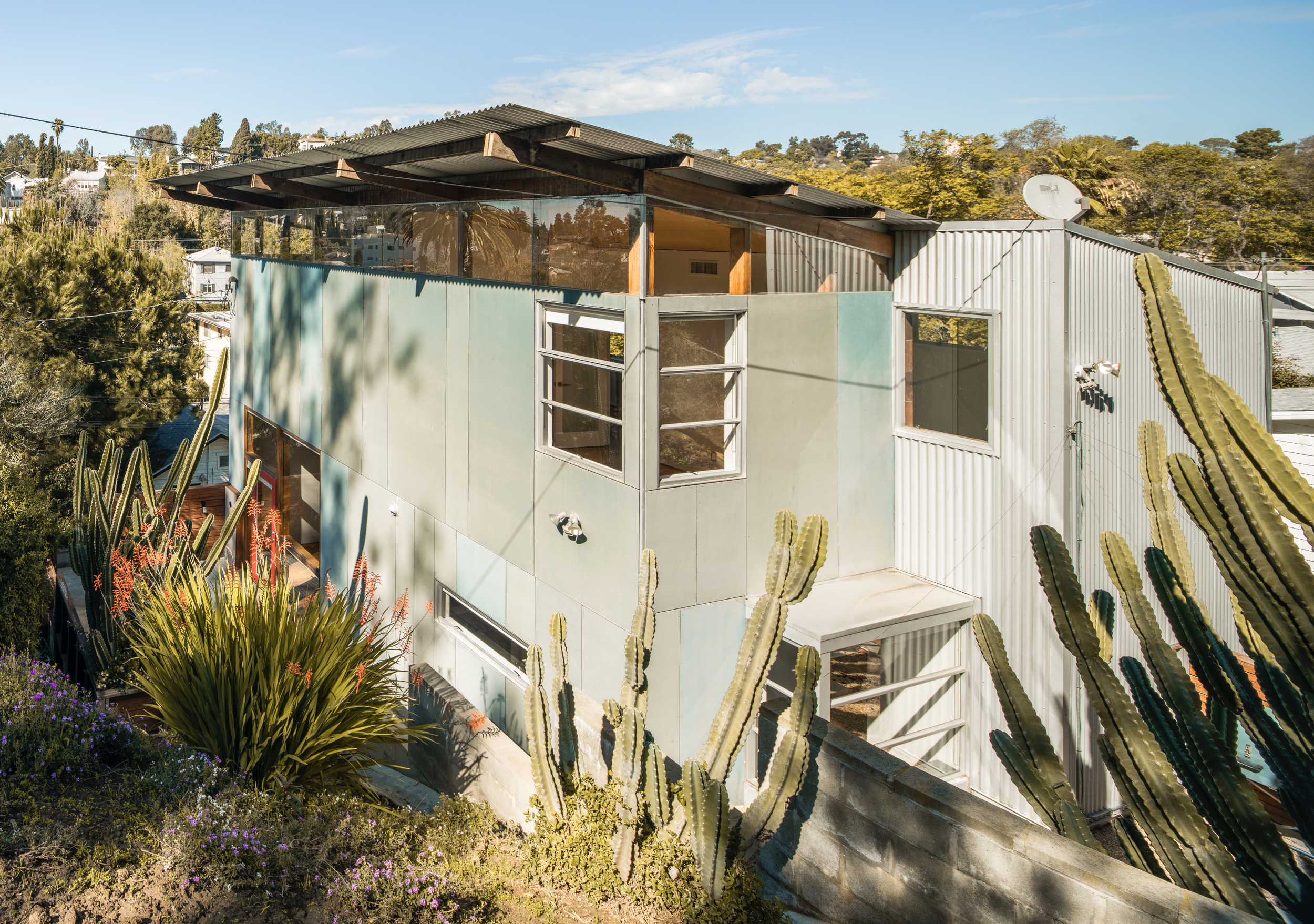 A modern two-story house with a mix of metal and pastel exterior walls, surrounded by desert plants including large cacti and flowering bushes, under a clear blue sky.