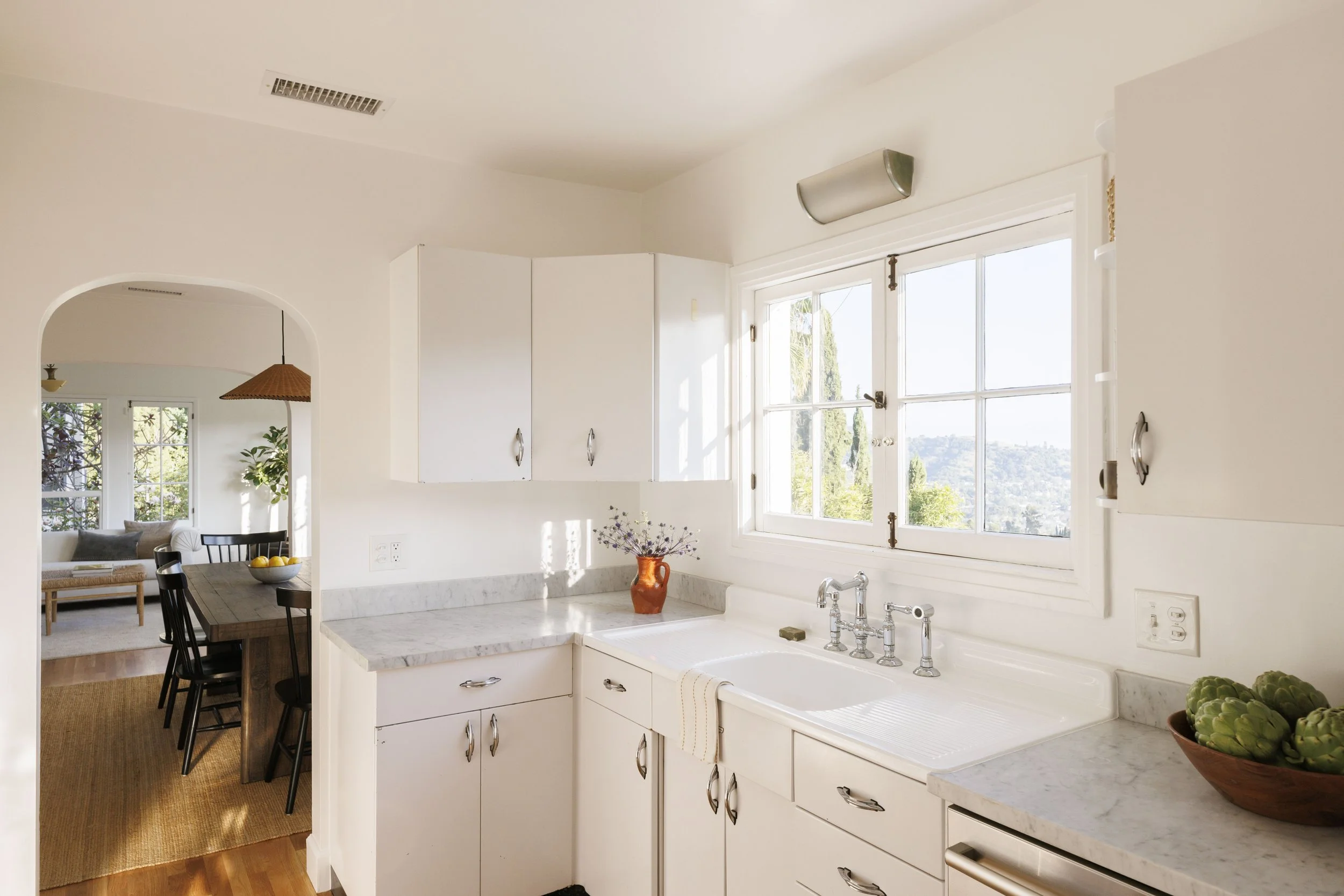 Bright kitchen with white cabinets, marble countertops, a large window above the sink, and a view of green trees outside.