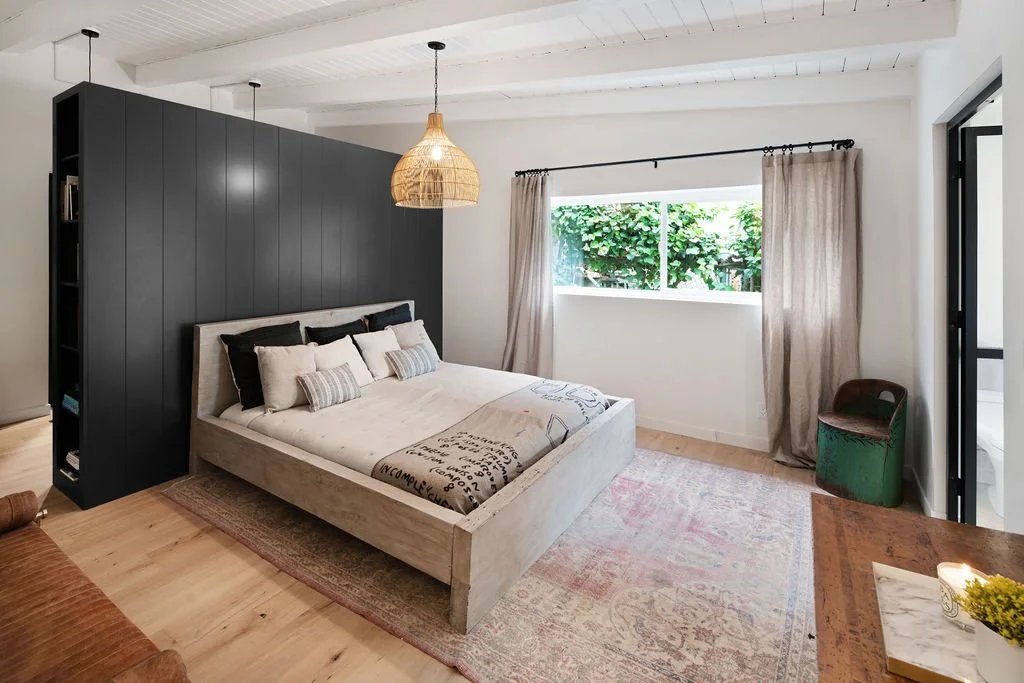 Modern bedroom with a bed, black headboard wall, wooden ceiling with pendant light, window with curtains, green chair, and wooden floor.
