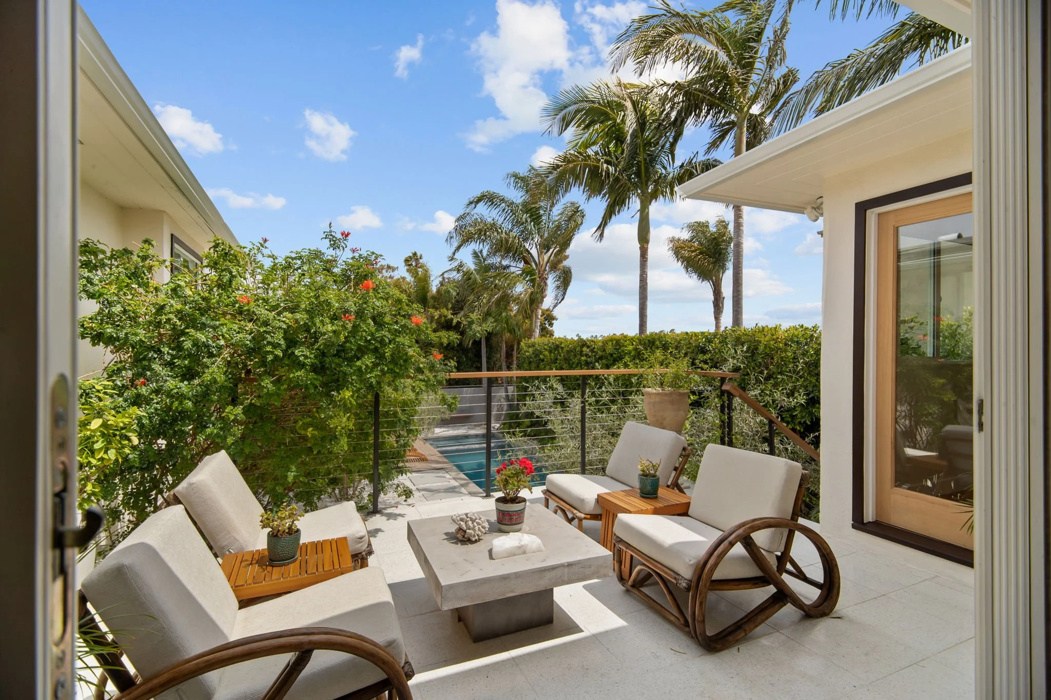 View of a balcony with outdoor seating, potted plants, palm trees, and a blue sky with clouds.